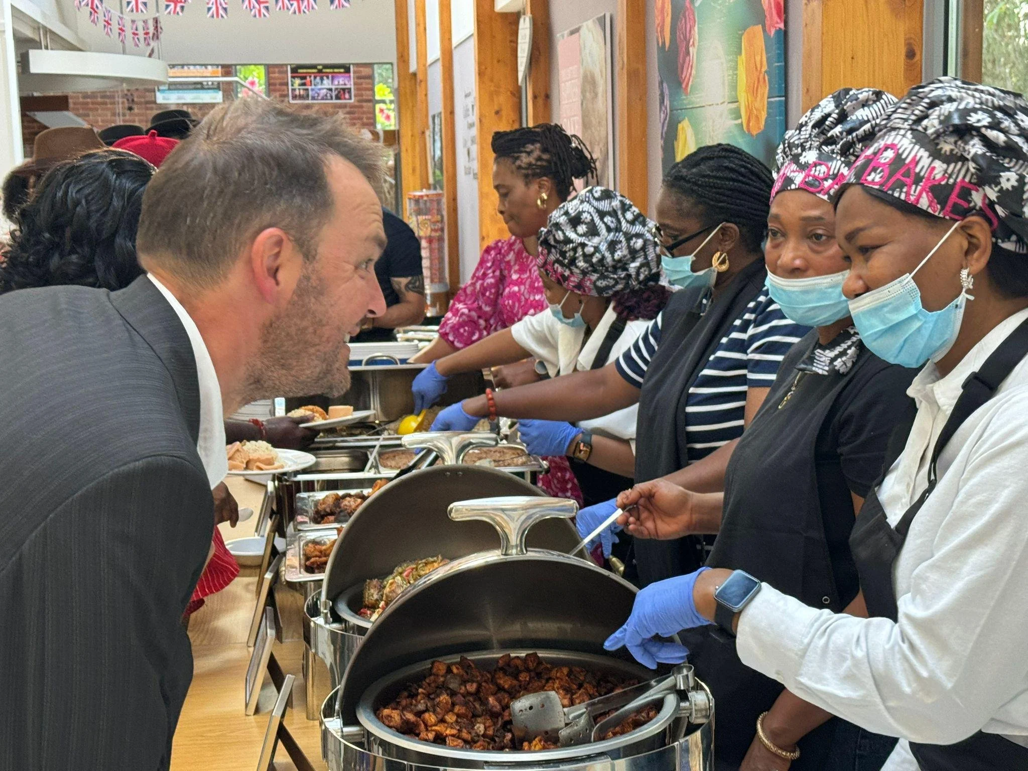 People wearing masks and gloves serving food at a buffet table, with a mix of men and women of diverse backgrounds, in a room decorated with bunting flags.