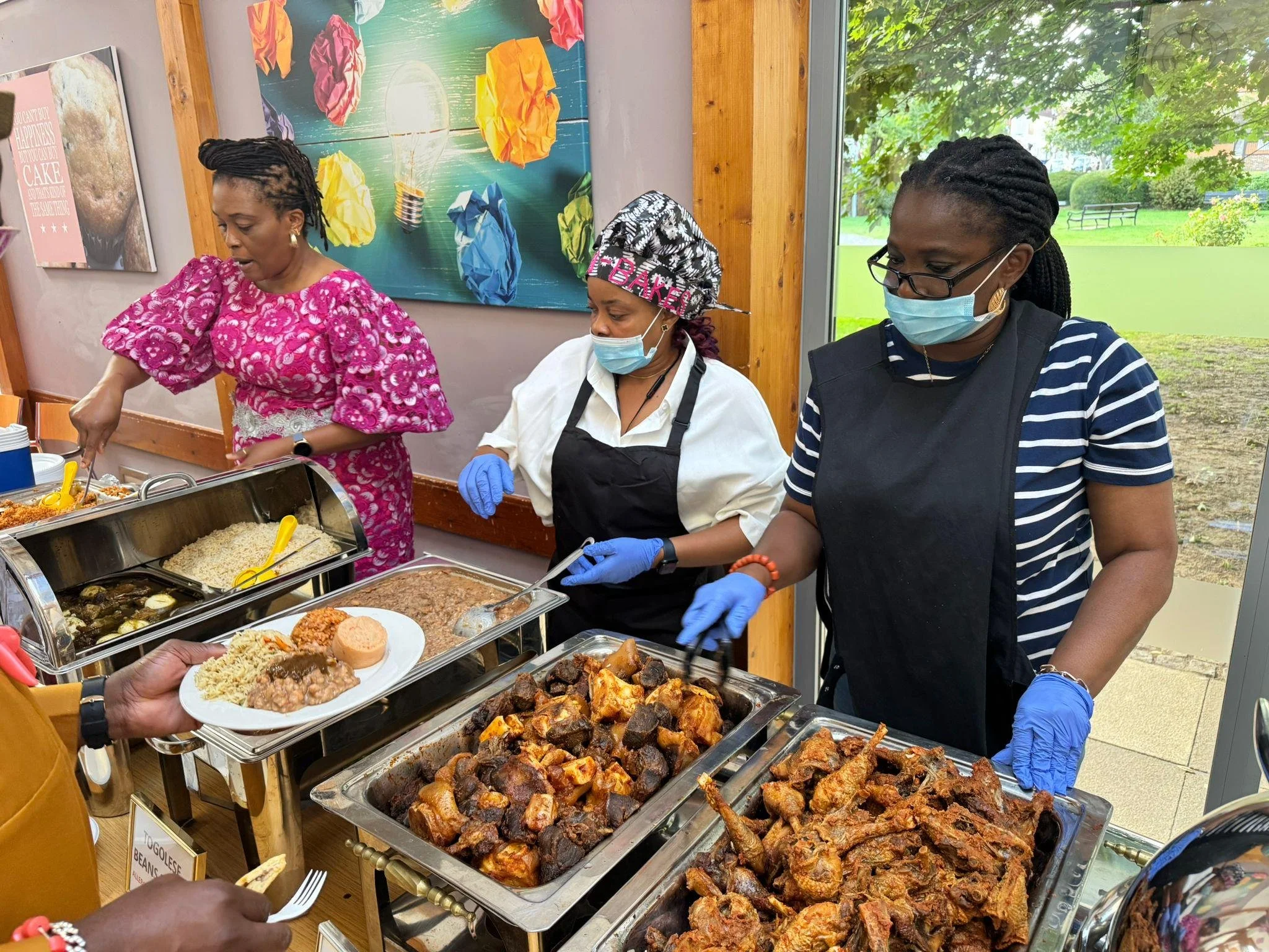 Three women wearing face masks and gloves serve food from trays at a buffet table indoors. The trays contain rice, cooked vegetables, meat, and fried items. Decorated walls and a large window with a view of greenery are in the background.