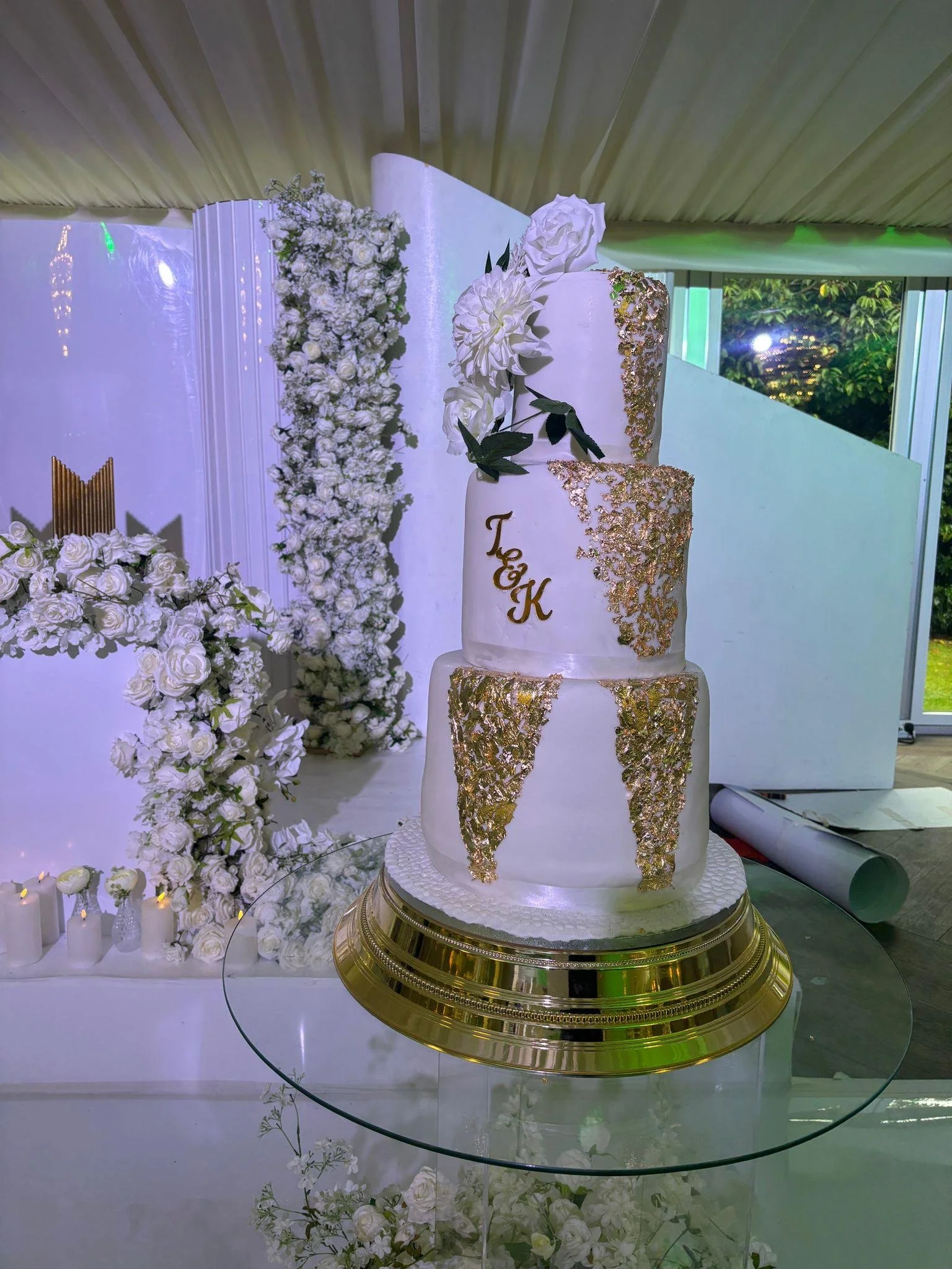 A four-tiered white wedding cake decorated with white flowers and gold accents, placed on a round golden cake stand, and surrounded by white floral arrangements and candles.