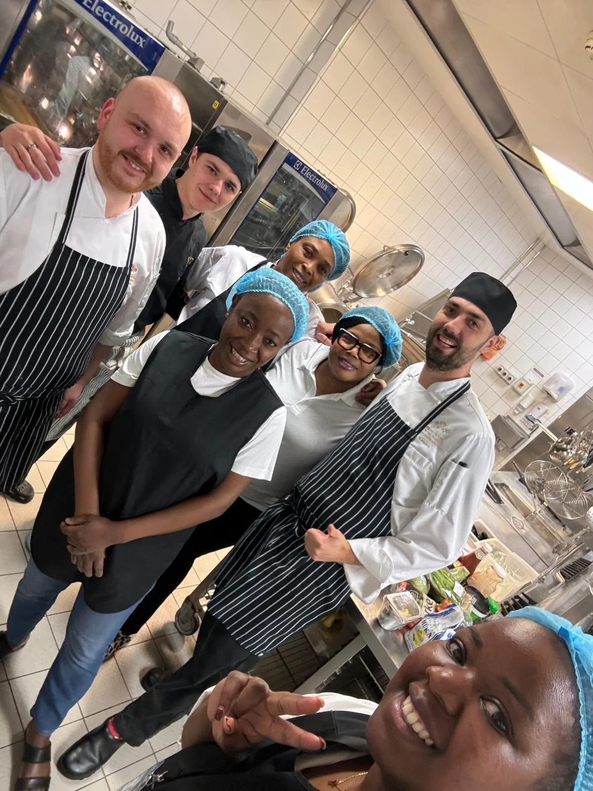 Group of six diverse chefs and kitchen staff in a commercial kitchen, smiling and posing for a selfie.