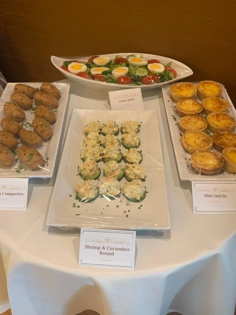 A buffet table with a green salad, shrimp and cucumber rounds, mini quiches, and chicken conquettes.