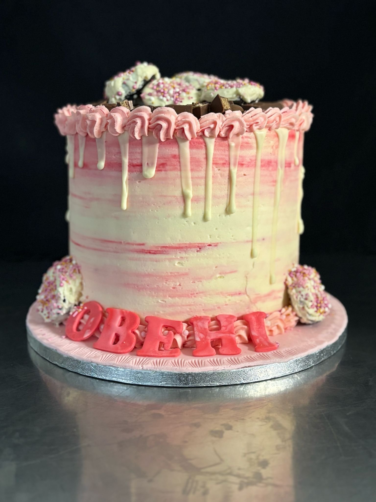 A pink ombre birthday cake decorated with pink and white icing, chocolate pieces, and sprinkles, with pink fondant letters spelling 'OBREH' on the cake board.