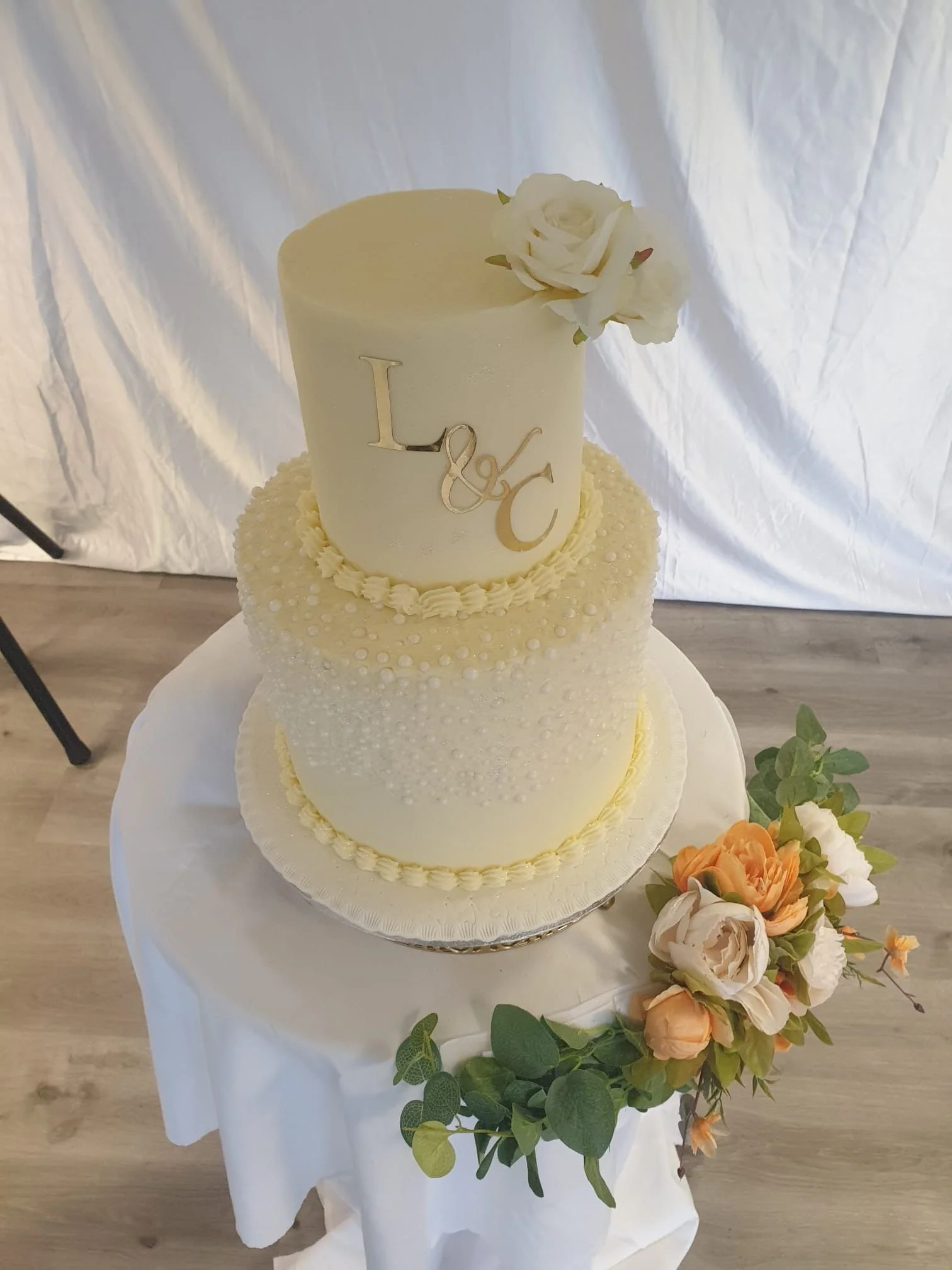 Cream-colored wedding cake with textured dots and floral topper, with a bouquet of peach, cream, and white flowers and greenery next to it on a white tablecloth.