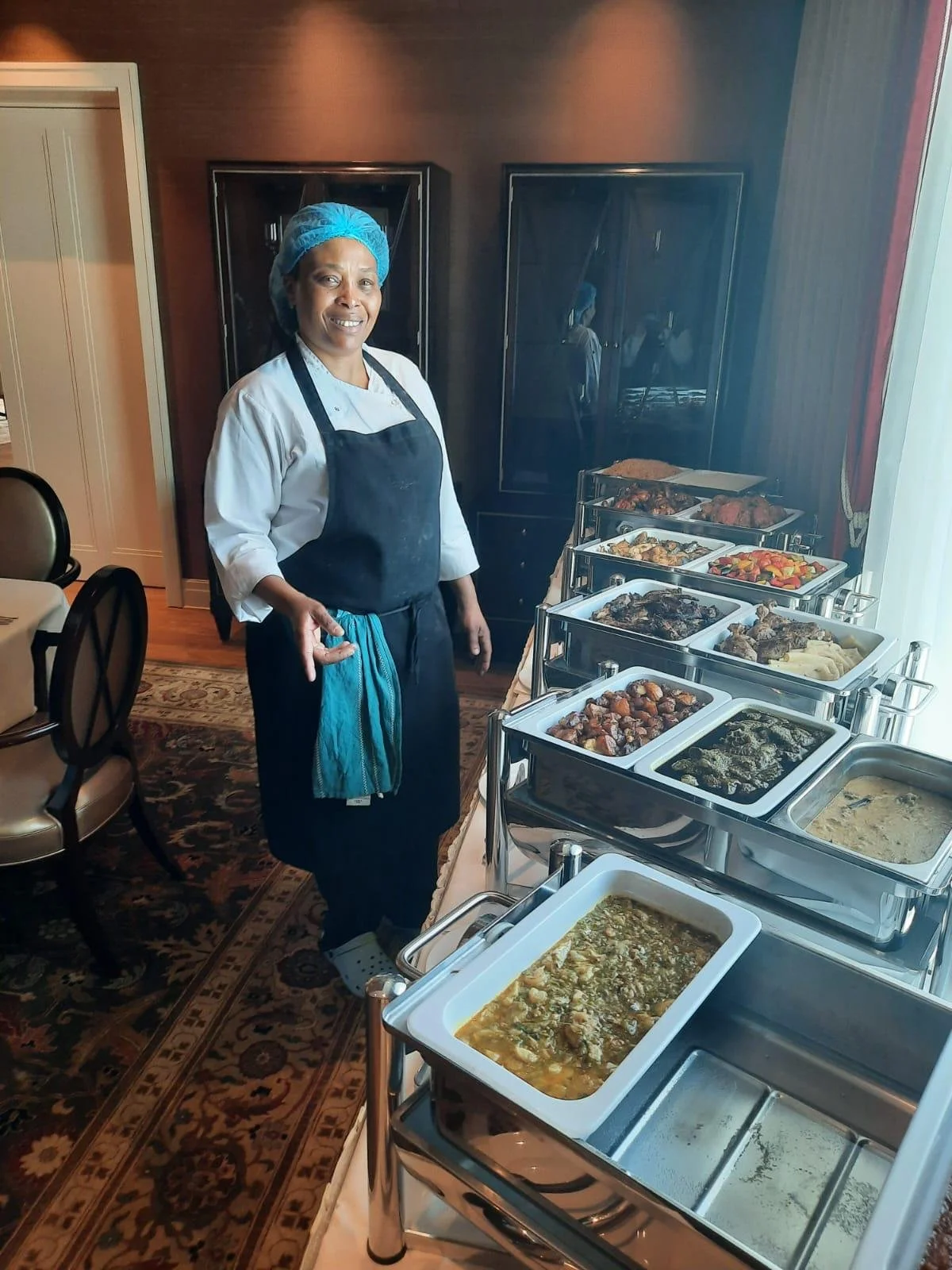 A woman in a chef's uniform and hairnet standing next to a buffet with various dishes in stainless steel chafing dishes. She is smiling and making an OK gesture with her hand.