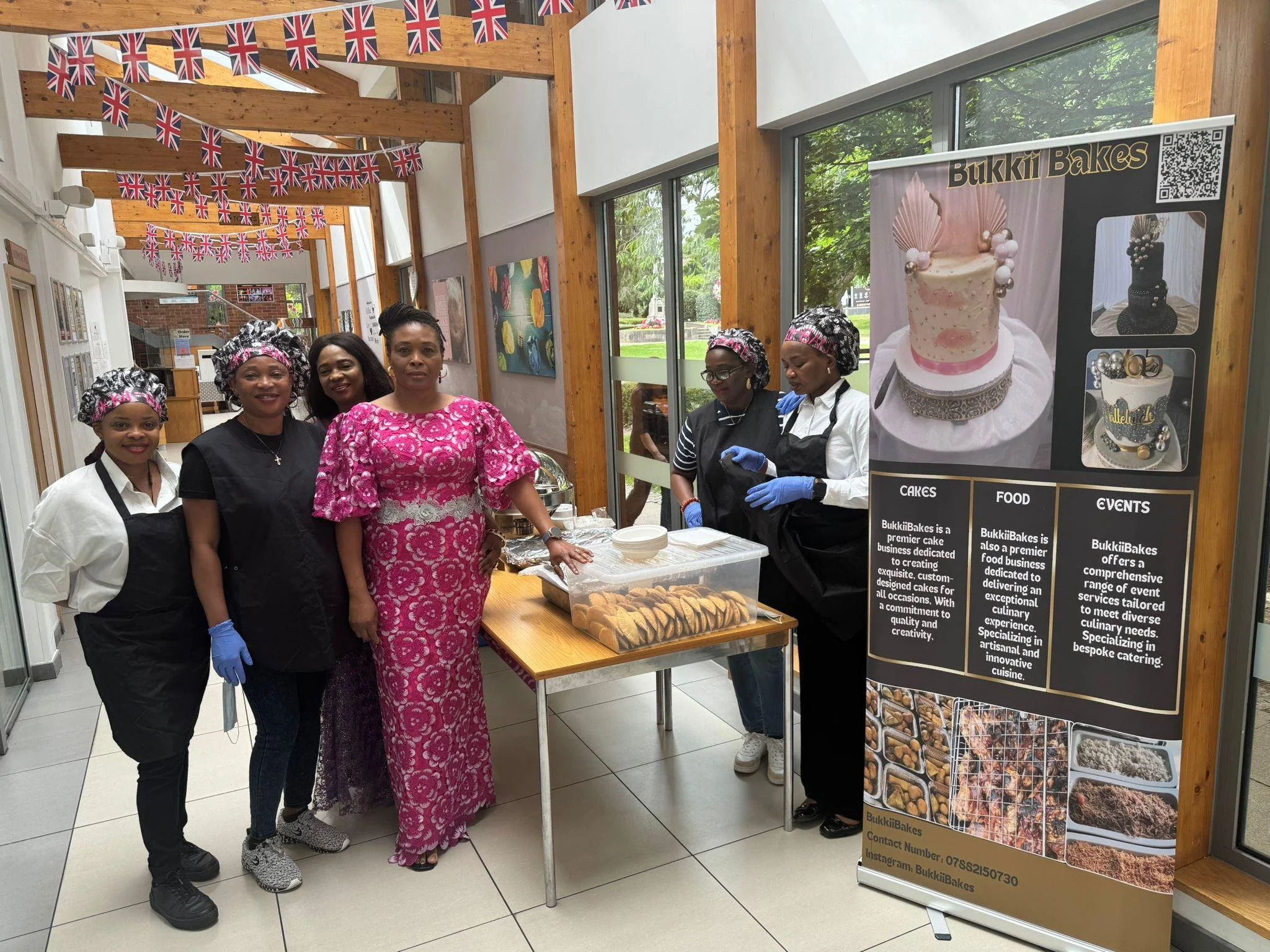 Group of five women standing behind a table filled with baked goods at an indoor event, with Union Jack flags hanging from the ceiling, and a large poster promoting Bukkii Bakes with images of decorated cakes.