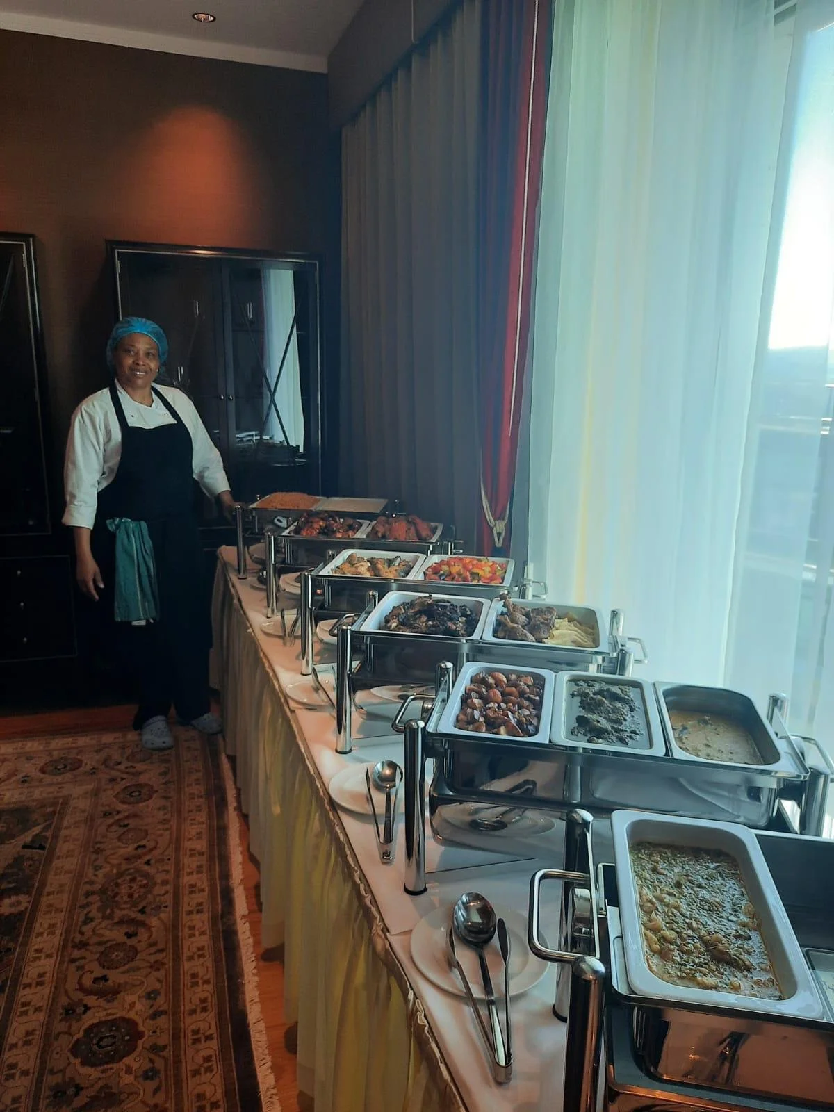 A chef standing next to a buffet table with various dishes in chafing dishes, in a room with a large window and curtains.