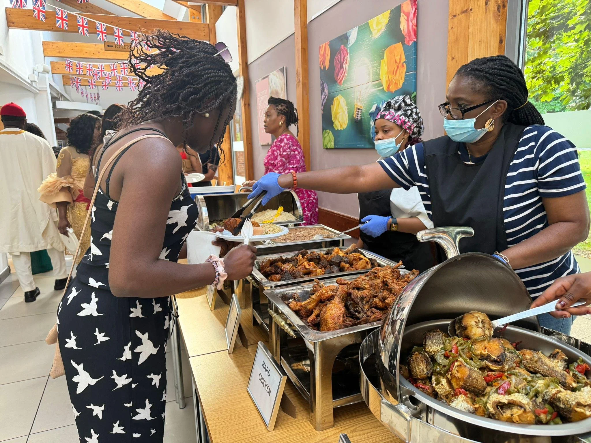 People wearing masks and gloves serving food at a buffet-style food station with various dishes, including roasted meats and vegetables, in a decorated indoor setting.