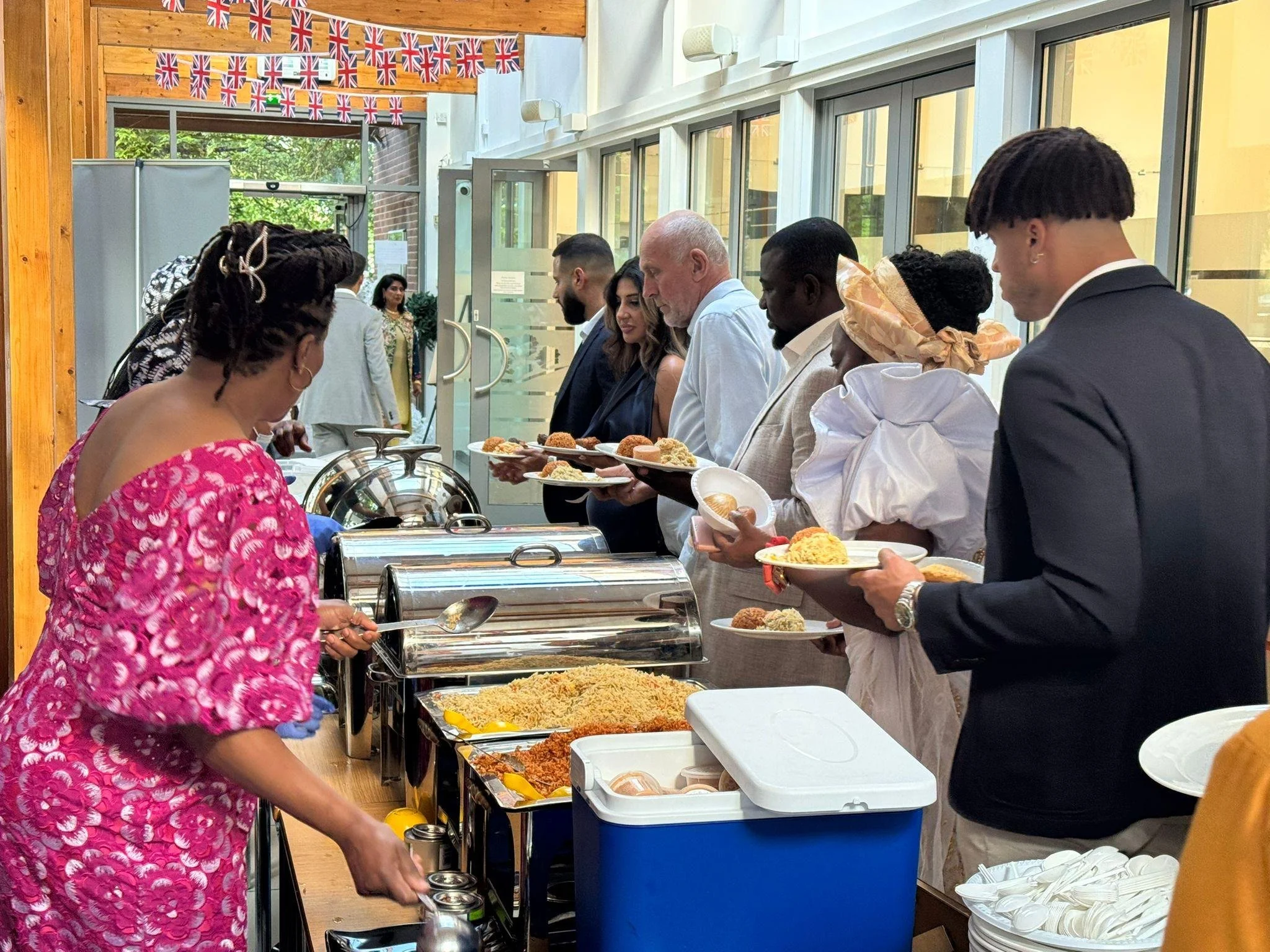 People in formal attire serving food at a buffet during a celebration decorated with Union Jack bunting.