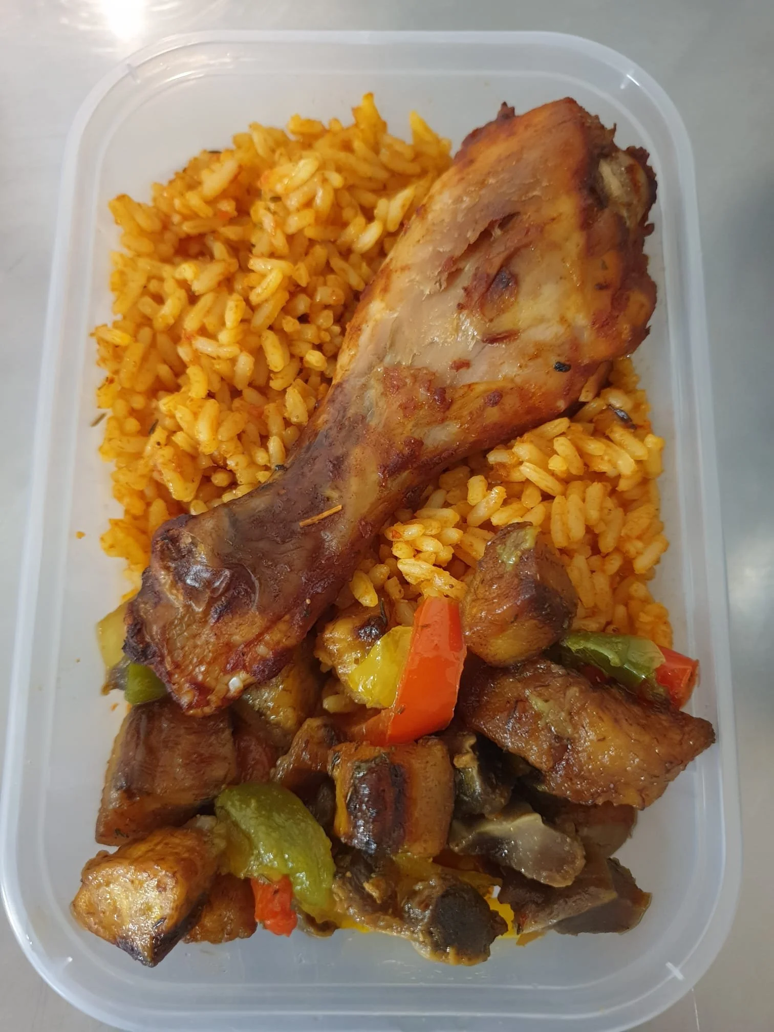 Cooked chicken drumsticks, seasoned, with visible vegetables and jollof rice in a plastic container.
