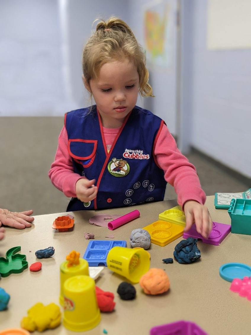 A young girl in a blue Awana vest, playing with colorful clay and cookie cutters at a table in a classroom.