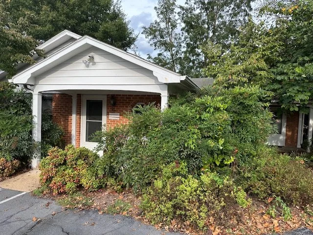 Front view of a small brick house with a gabled roof, surrounded by overgrown bushes and plants, with a parking lot in the foreground and cloudy sky above.