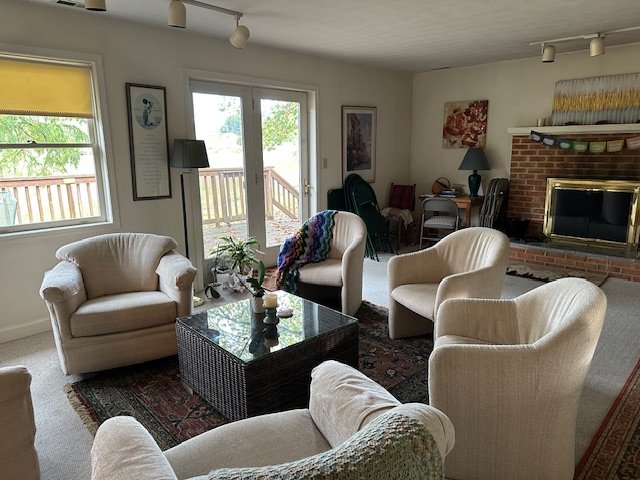 Living room with cream-colored armchairs, a black coffee table, and a brick fireplace. Large windows and a sliding glass door lead outside, with a deck and trees visible. Decor includes framed art, a lamp, and a blanket on one chair.