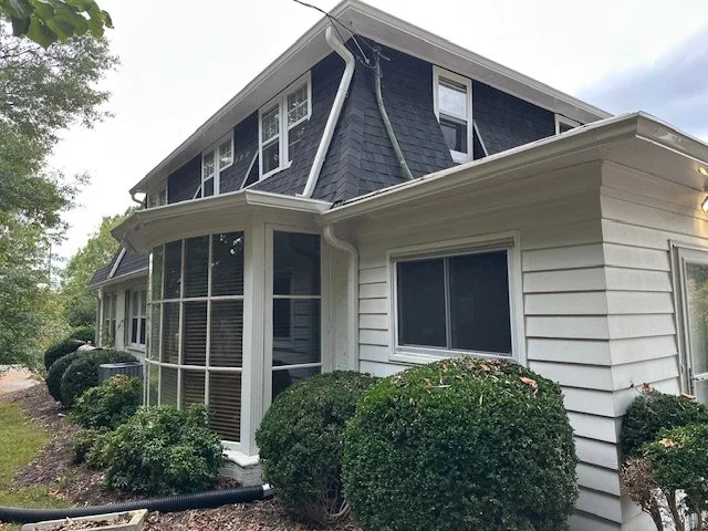 Exterior view of a house with a two-story section and a single-story sunroom, surrounded by shrubbery and trees.