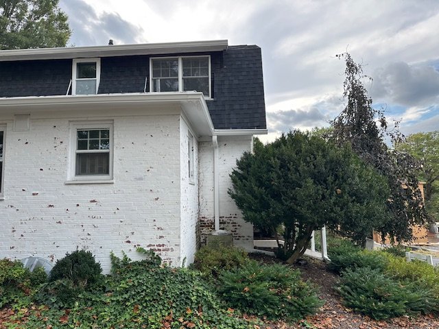 A white two-story house with black shingles on the upper level and a small front yard with green shrubs and a tree, under a cloudy sky.