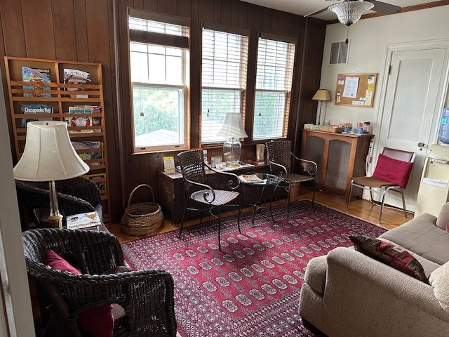 A cozy room with wooden walls and large windows, furnished with wicker chairs, a beige sofa, a red-patterned rug, and a small table. Cozy lamps, a bookshelf, and a bulletin board decorate the space.