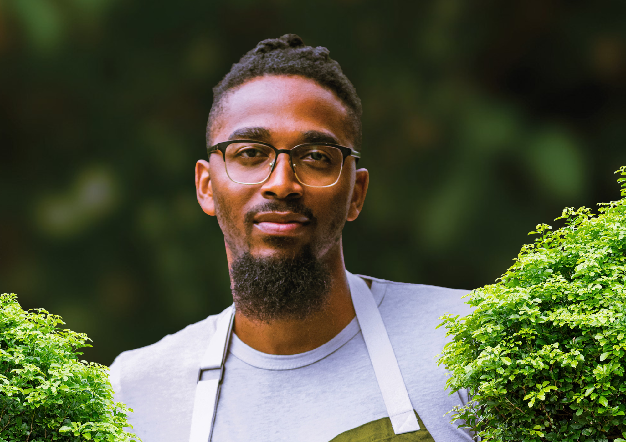 A man with glasses and a beard stands outdoors near green bushes, with dark blurred background, wearing a light gray t-shirt.