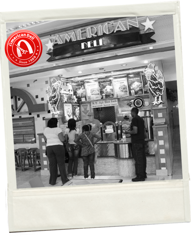 People standing in line at a fast food restaurant named American Deli inside a shopping mall.