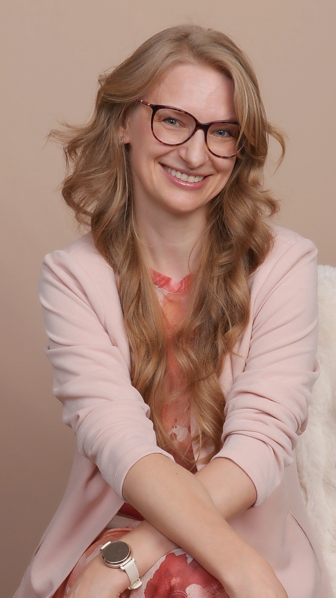 A woman with long, wavy red hair and glasses, smiling with her arms crossed, sitting against a neutral background.