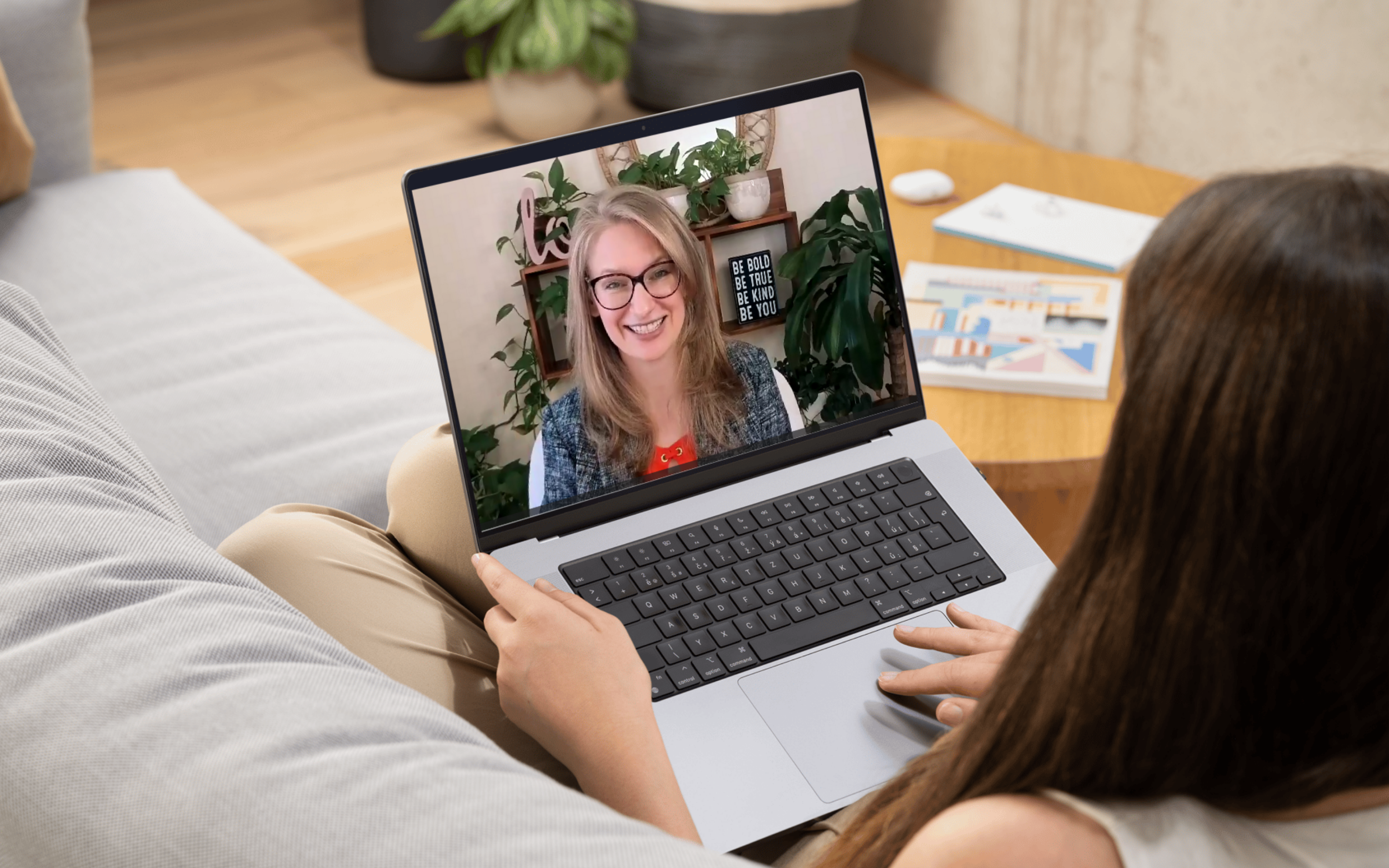 Person with long brown hair waving at a woman on a video call on a laptop, sitting on a beige couch with a plant and books in the background.