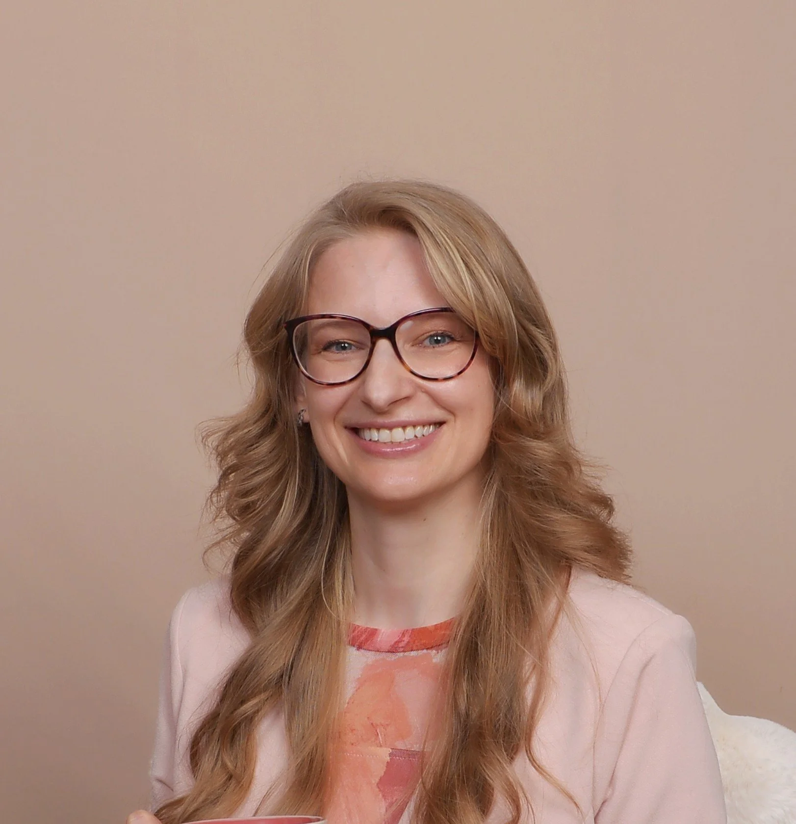 Portrait of a woman with wavy, blonde hair and glasses, smiling against a neutral background.