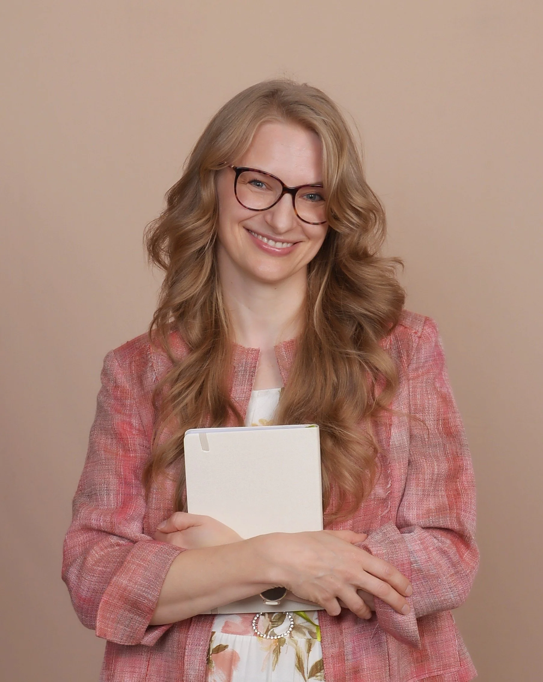 A woman with long, curly red hair, glasses, and a pink blazer holding a tablet, standing against a beige background.