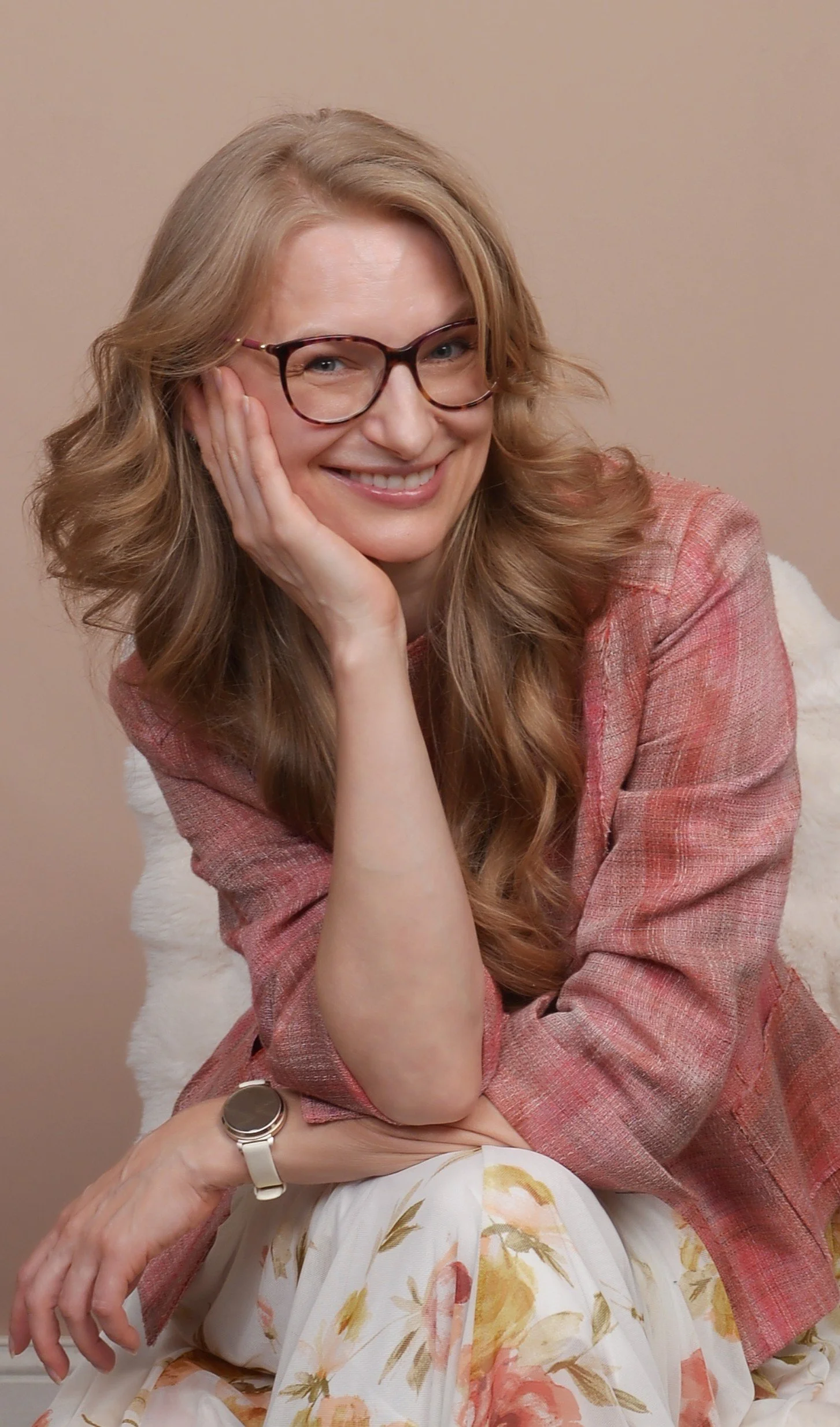 A woman with long wavy blonde hair, glasses, and a warm smile, sitting with her hand resting on her cheek. She is wearing a pinkish-red blazer and a floral skirt, and a watch on her wrist. The background is a plain beige wall.