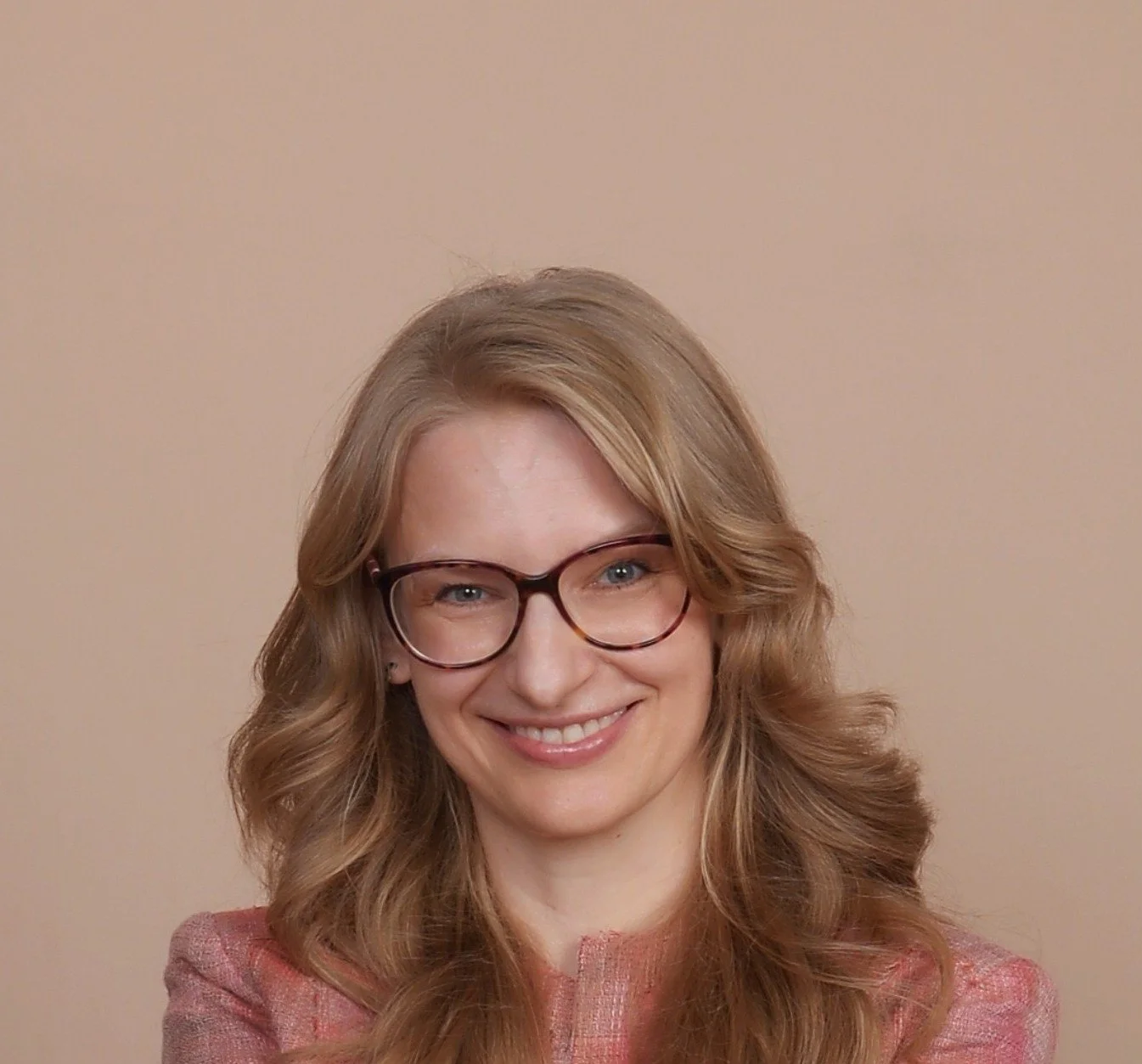A woman with long, wavy blonde hair, glasses, and a pinkish-red blazer smiling against a beige background.