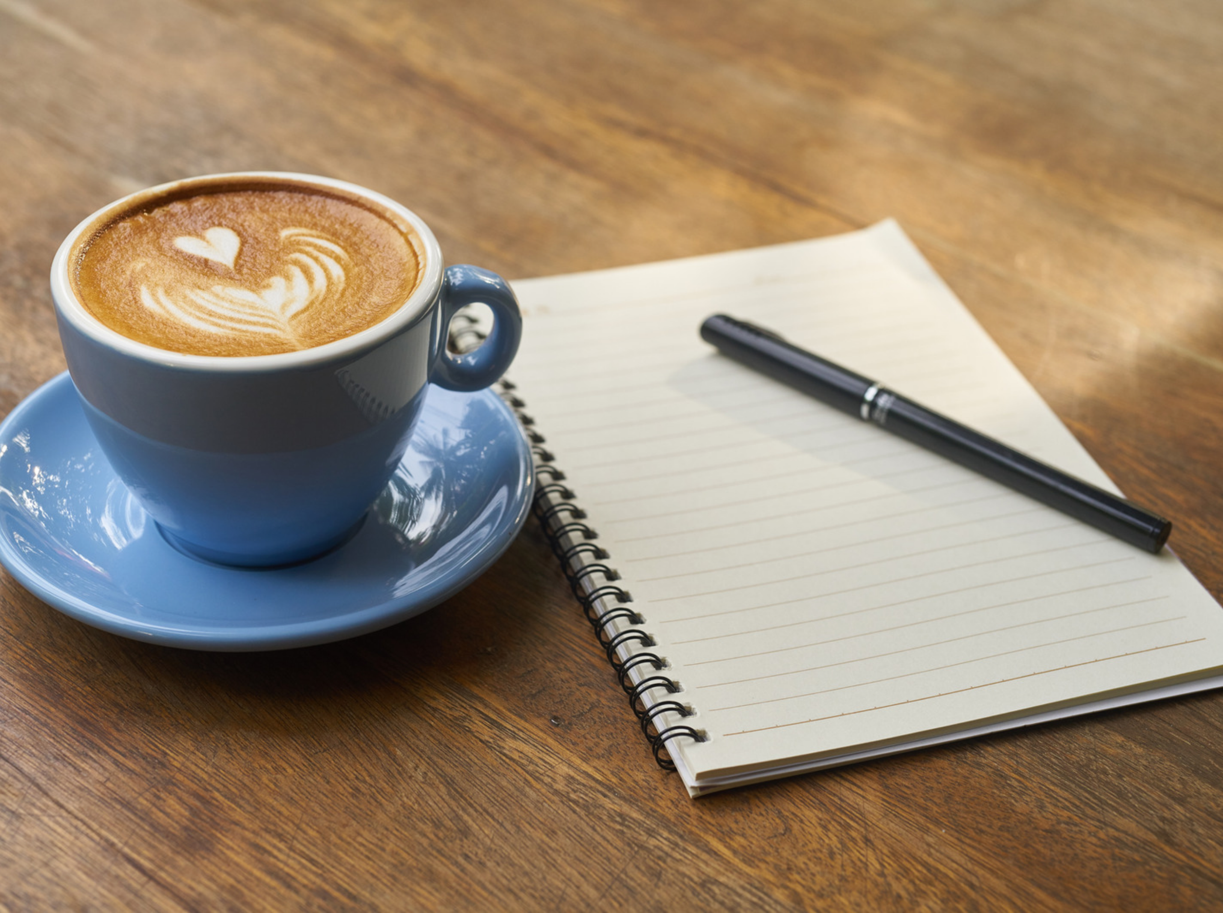 A blue cup of coffee with latte art in a heart pattern sits on a matching blue saucer on a wooden table. Next to the cup, there is a spiral notebook with blank lined paper and a black pen resting on top of it.