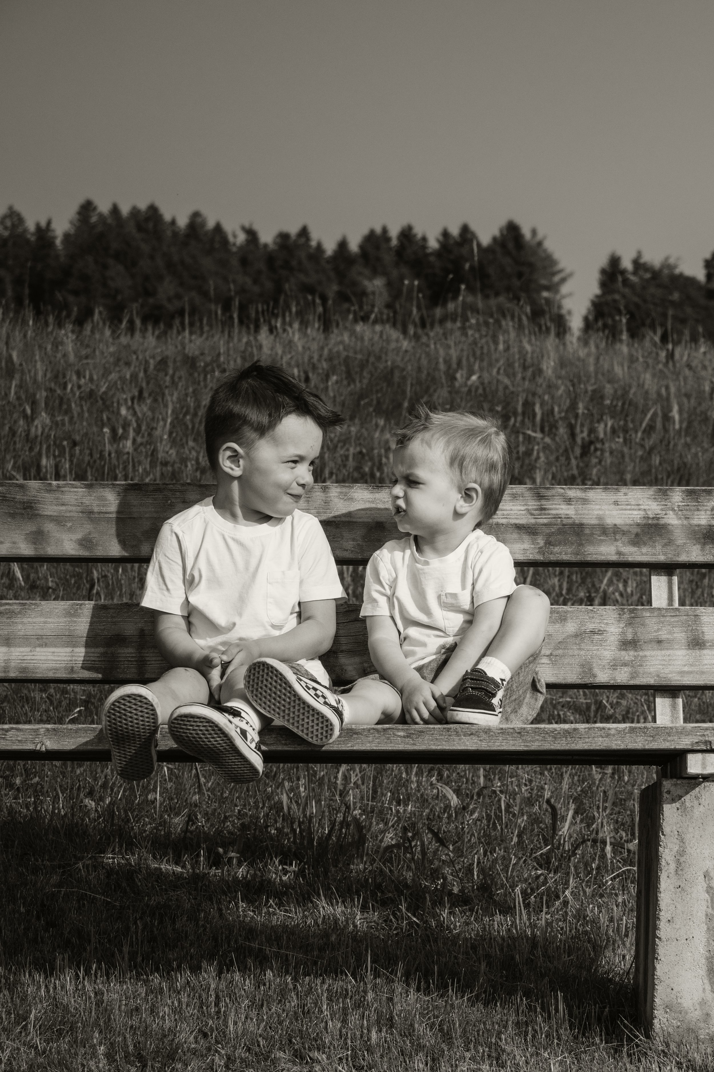 Zwei Kinder sitzen auf einer Parkbank und schauen sich an, im Hintergrund Bäume und Gras, Schwarz-Weiß-Foto.