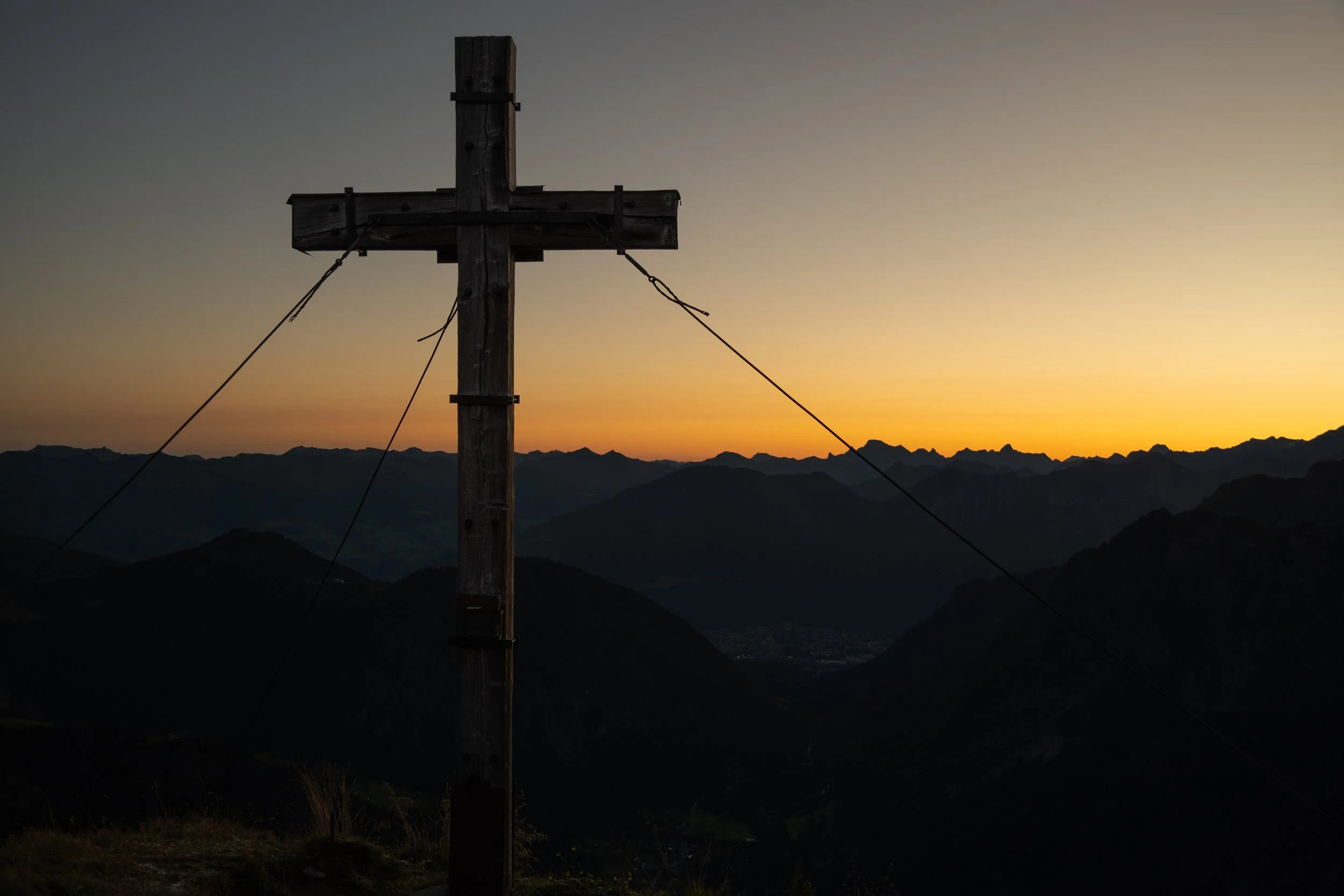 Ein Holzkreuz auf einem Berg bei Sonnenaufgang, umgeben von Bergketten im Hintergrund.