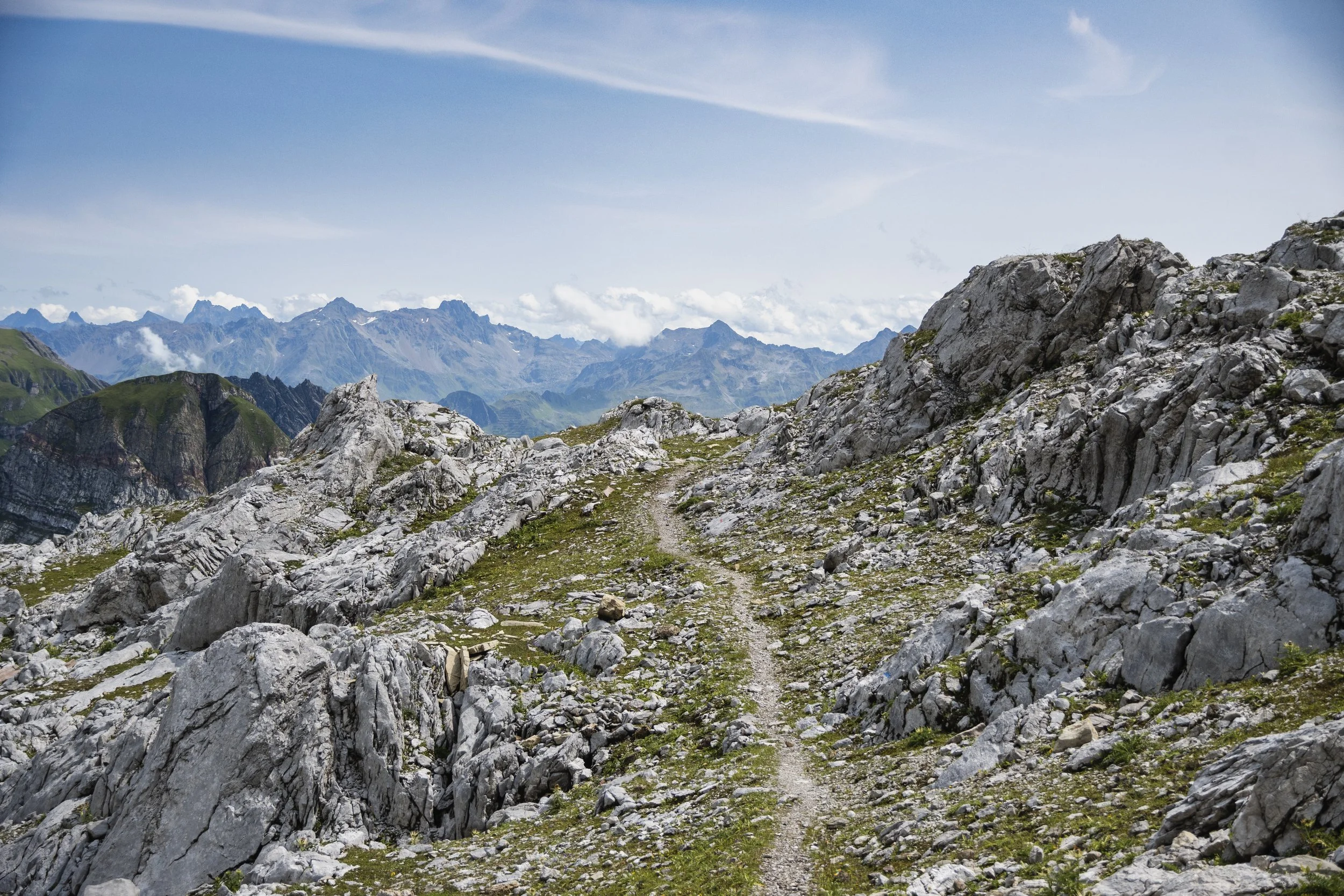 Ein schmaler Wanderweg durchquert eine felsige Gebirgslandschaft mit hohen Berggipfeln im Hintergrund, unter einem klaren blauen Himmel.