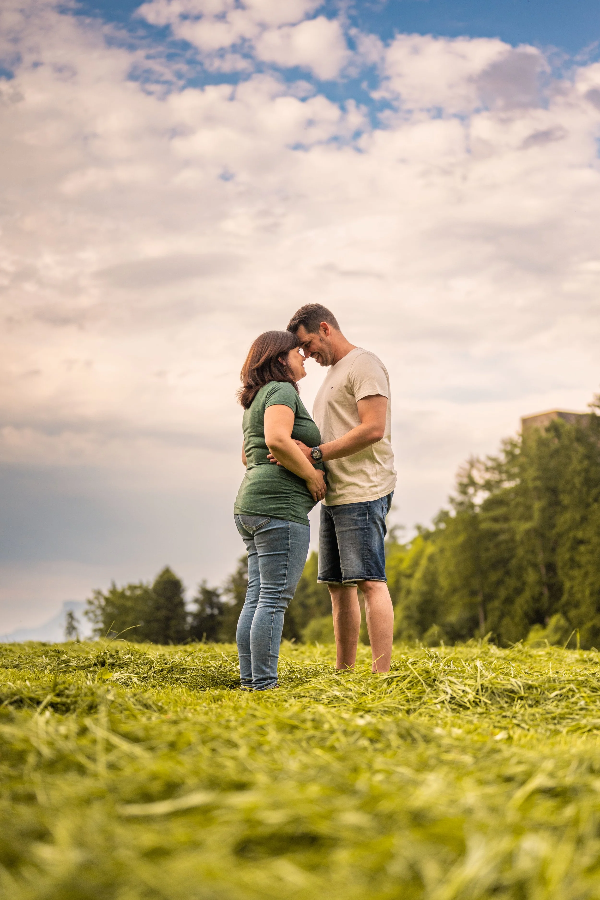 Auf frisch geschnittenem Gras mit schöner Wolkenstimmung stehen sich eine schwangere Frau und ihr Mann gegenüber. Mit den Köpfen aneinander halten sie gemeinsam den wunderschönen Babybauch.