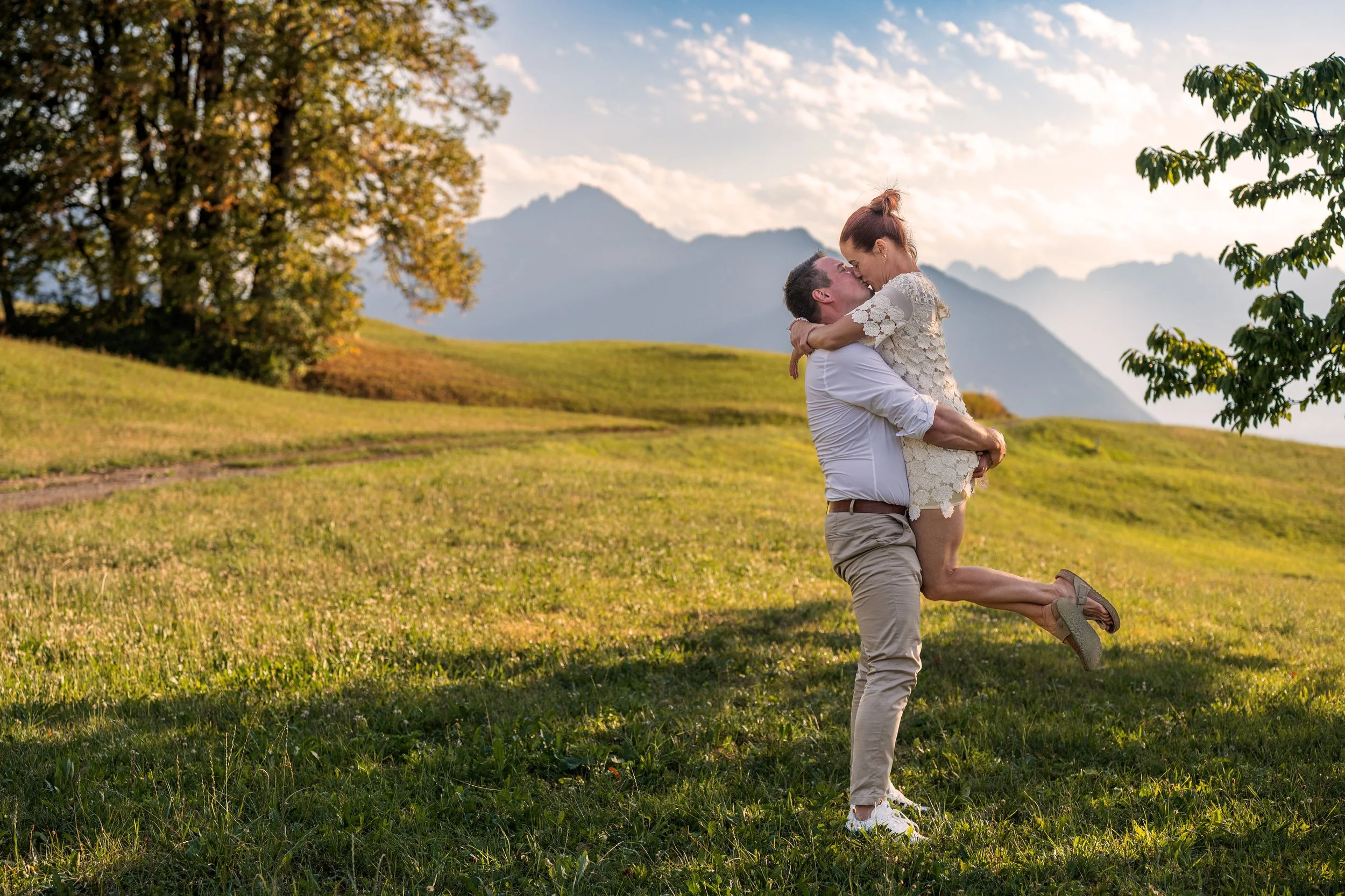 Ein Mann hebt seine Frau hoch. Sie umarmen und küssen sich. Im Hintergrund eine schöne Bergkulisse mit Wolkenstimmung.