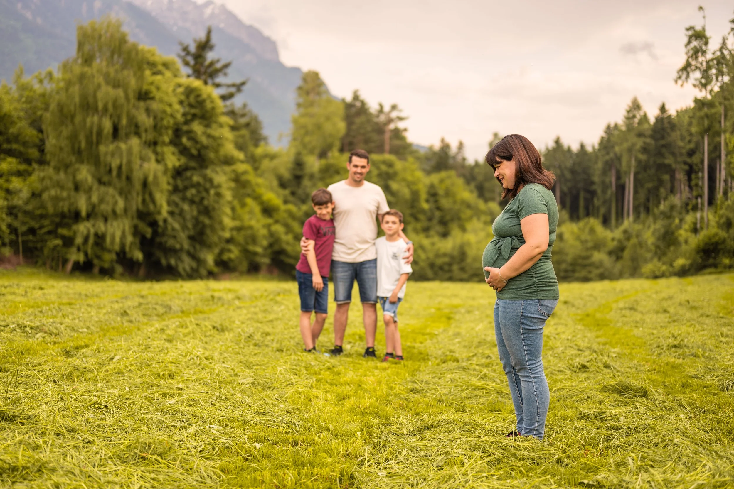 Die schwangere Frau im Fokus, sie hält ihren Babybauch und blickt lächelnd nach unten. Weiter hinten der Rest der Familie - ihr Mann und die beiden Söhne - Blick Richtung Mama.