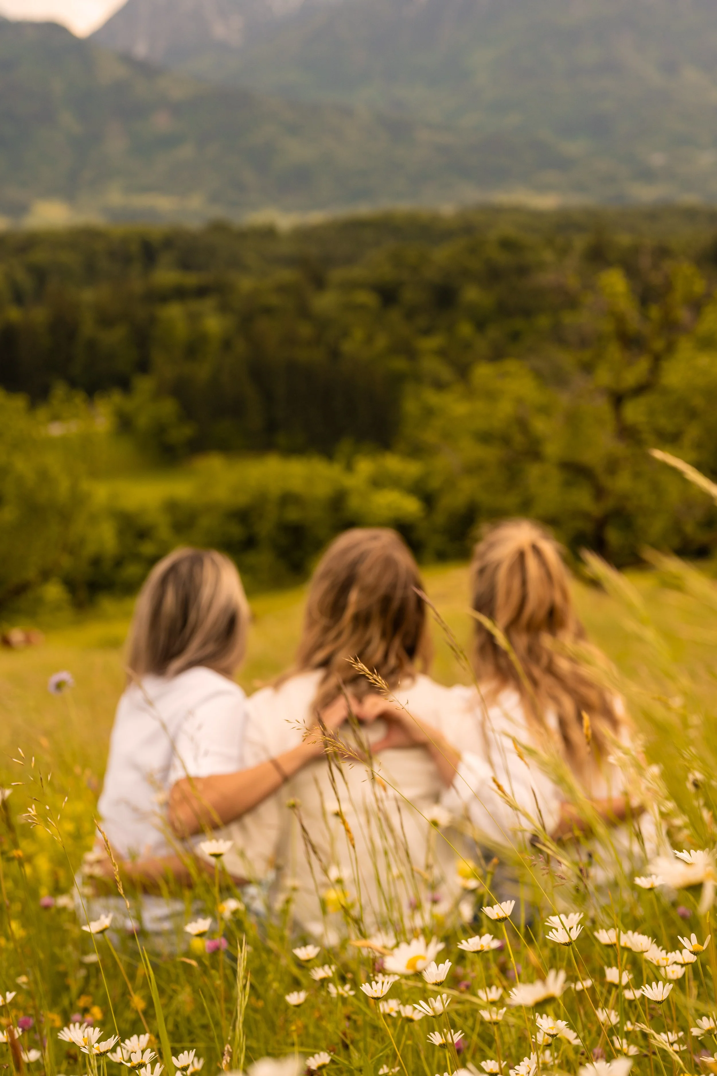 Mit dem Rücken zur Kamera blicken drei Frauen in Richtung Berge. Sitzend in einer Blumenwiese formen sie mit den Händen ein Herz auf dem Rücken der in der Mitte sitzenden Frau.