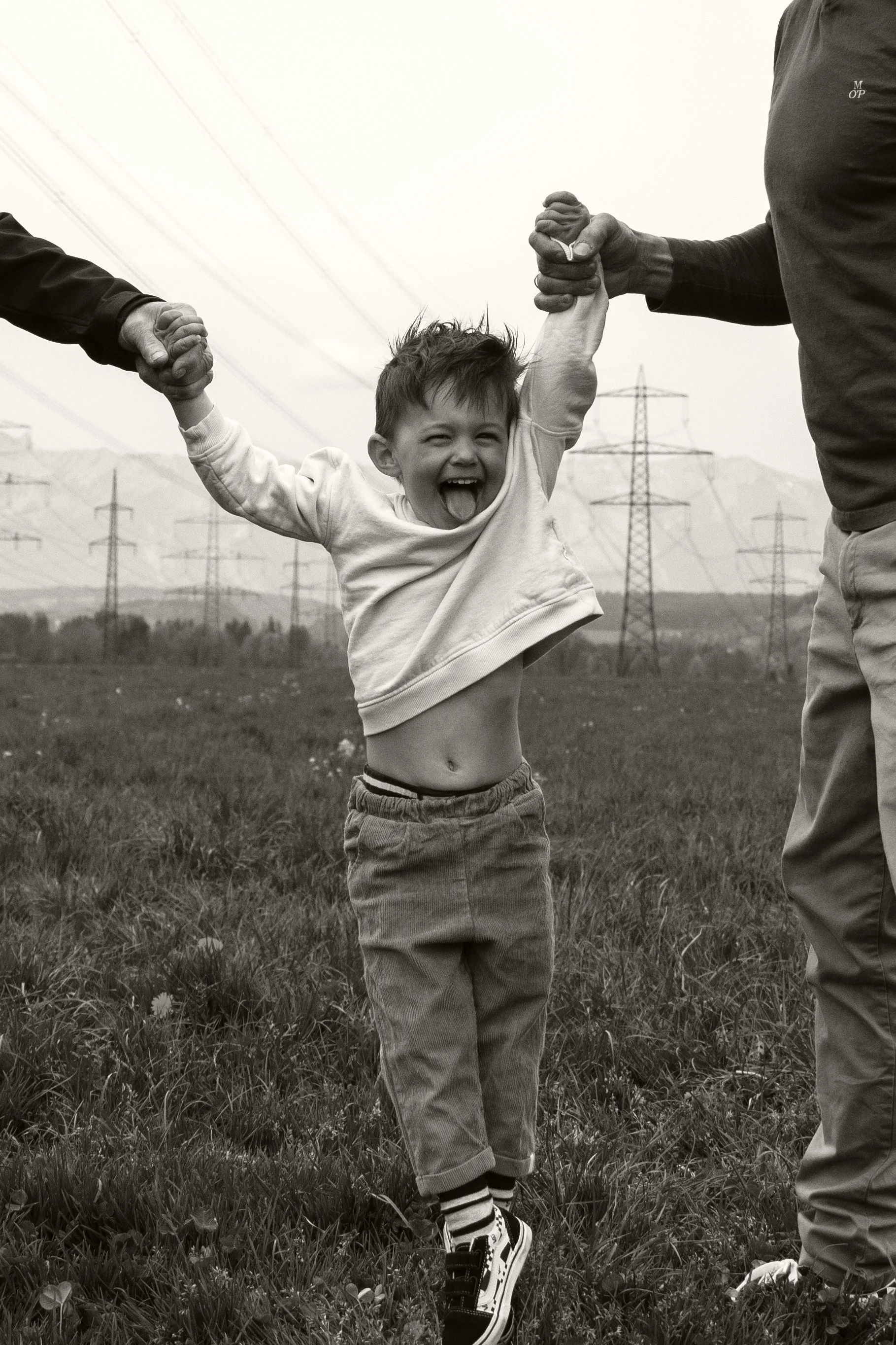 Ein lachendes Kind hängt an den Händen von zwei Erwachsenen in einem offenen Feld, im Hintergrund Stromleitungen und Berge, schwarz-weiß.
