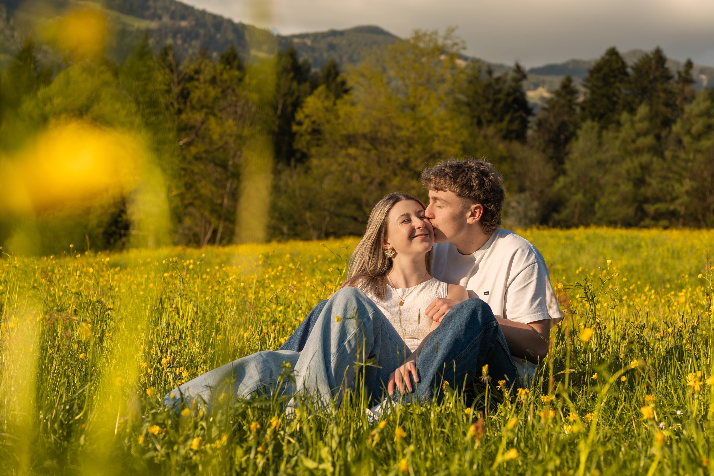 Ein romantisches Paarfoto, sitzend auf einer gelben Blumenwiese geben sich die beiden einen Kuss auf die Wange.