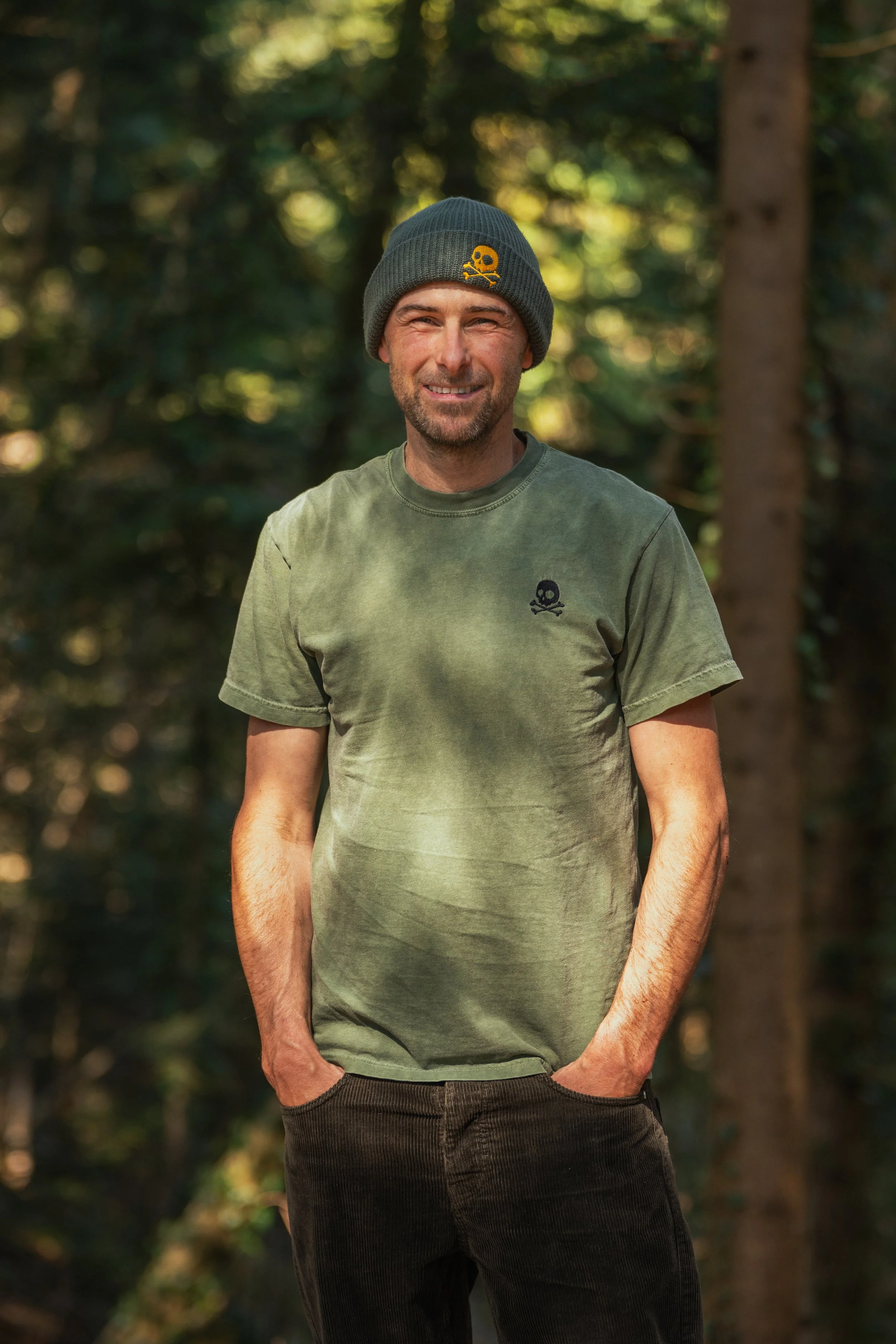 Ein Mann in grünem T-Shirt und grüner Mütze steht lächelnd im Wald.