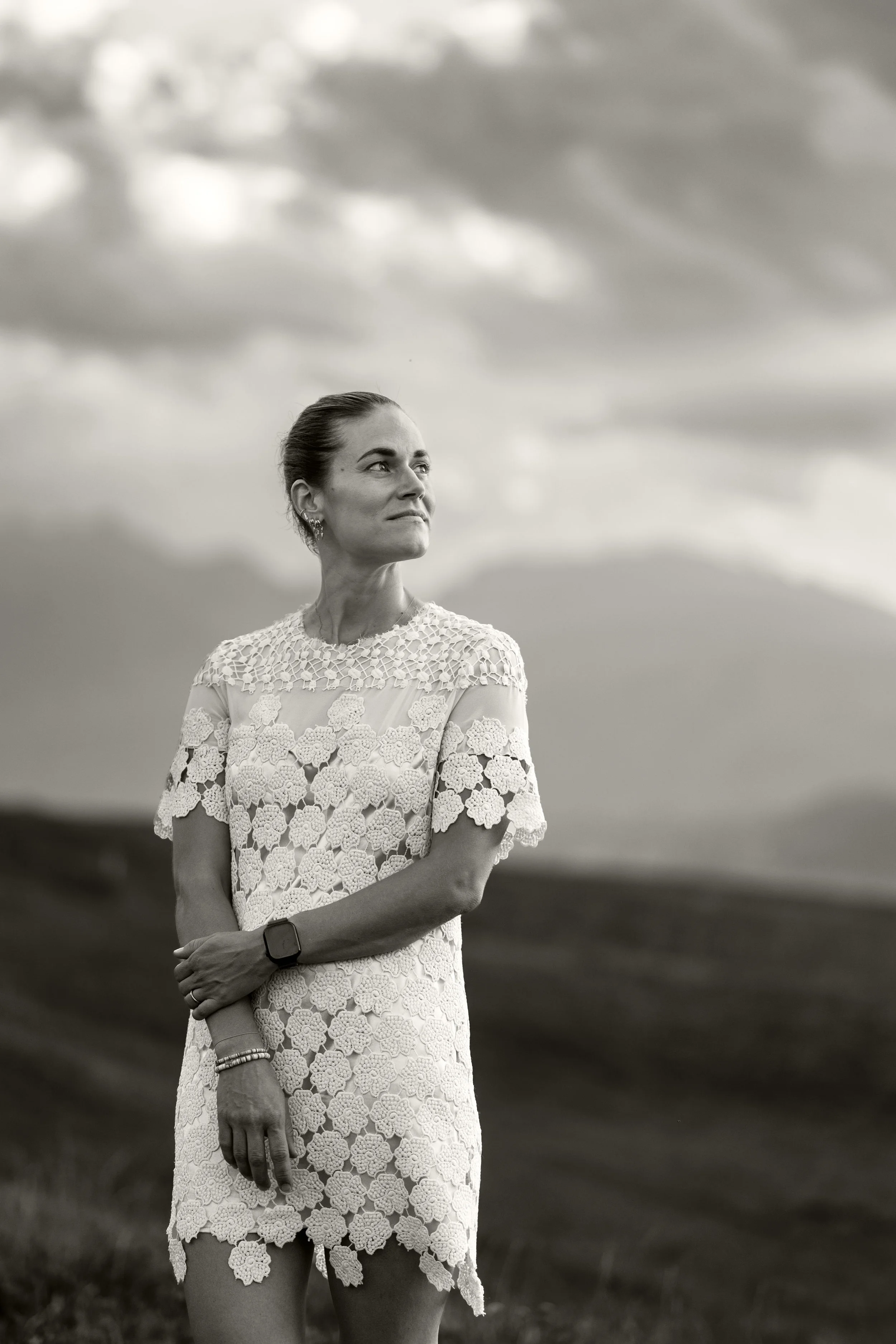Ein schwarz-weiß Foto einer Frau. Sie blickt hoch zum Himmel und hält sich mit der einen Hand an ihrem Arm. Sie trägt ein weißes Spitzenkleid. Im Hintergrund eine schöne Abendstimmung. 