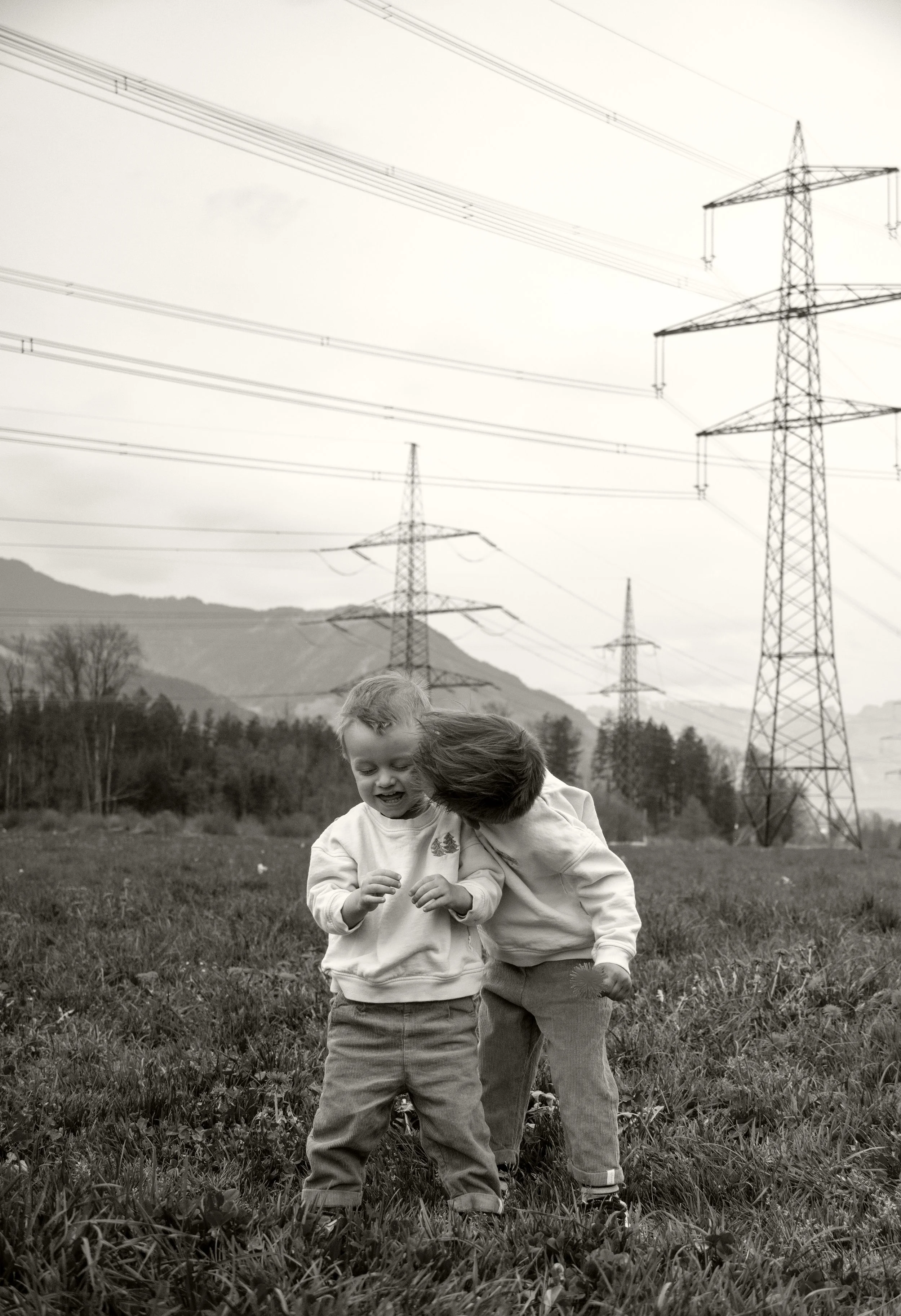 Zwei Jungen stehen in einem grasbewachsenen Feld mit Hochspannungsleitungen im Hintergrund. Einer der beiden gibt dem anderen einen Kuss.