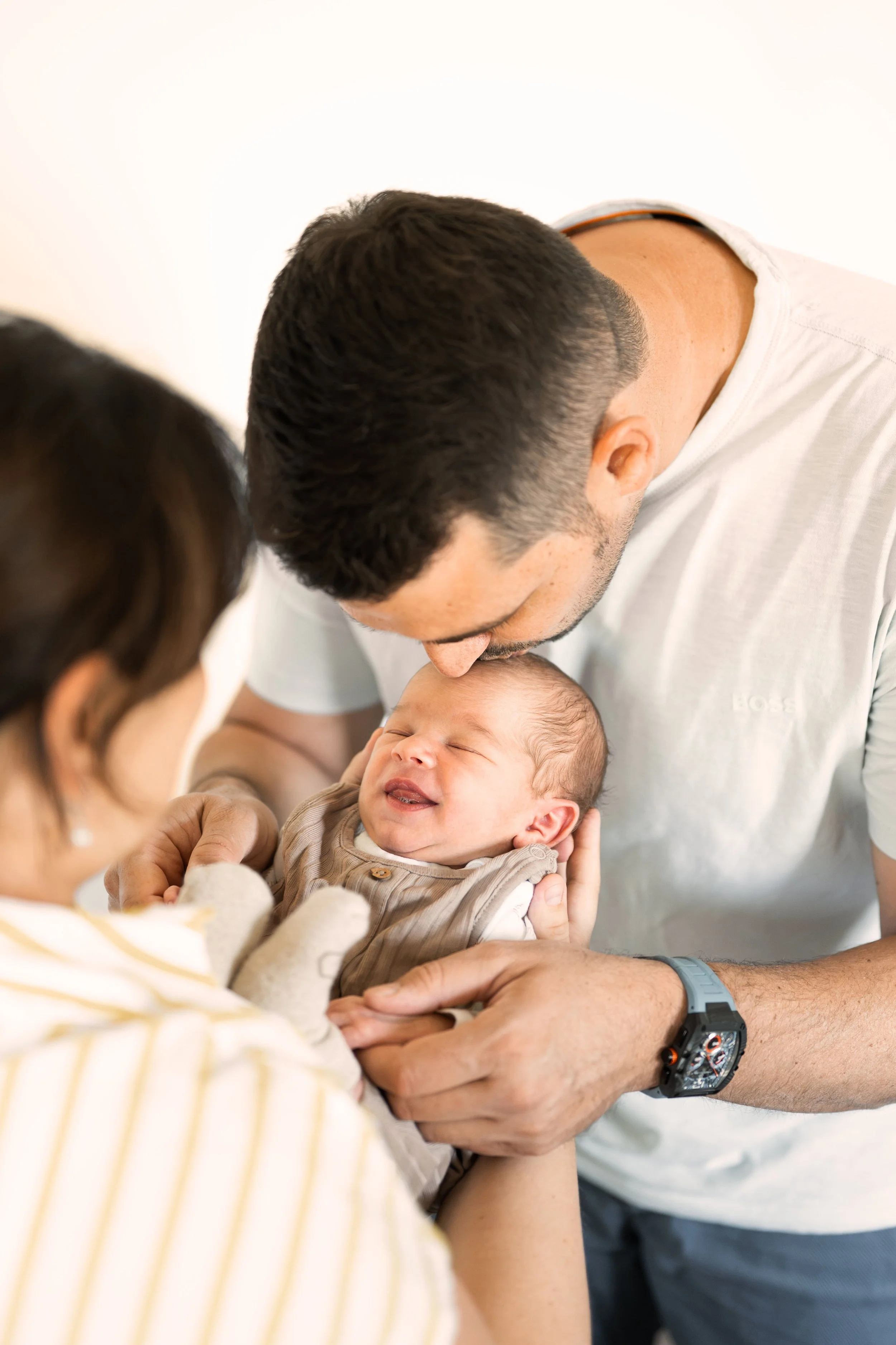 Ein Baby liegt geborgen in den Händen seiner Mutter. Gegenüber der Vater. Er beugt sich über das Kind und gibt ihm liebevoll einen Kuss auf die Stirn, dabei hält er sanft die Hände des Babys.
