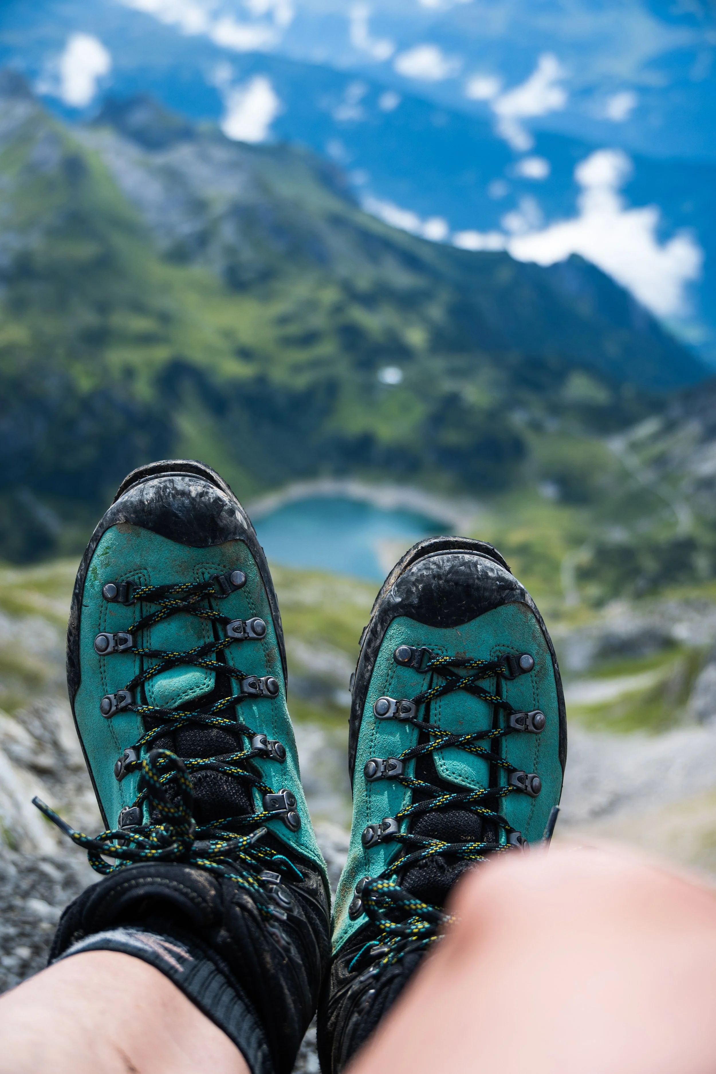 Bergschuhe von oben fotografiert. Im Hintergrund ein See umgeben von Bergen und weißen Wolken.