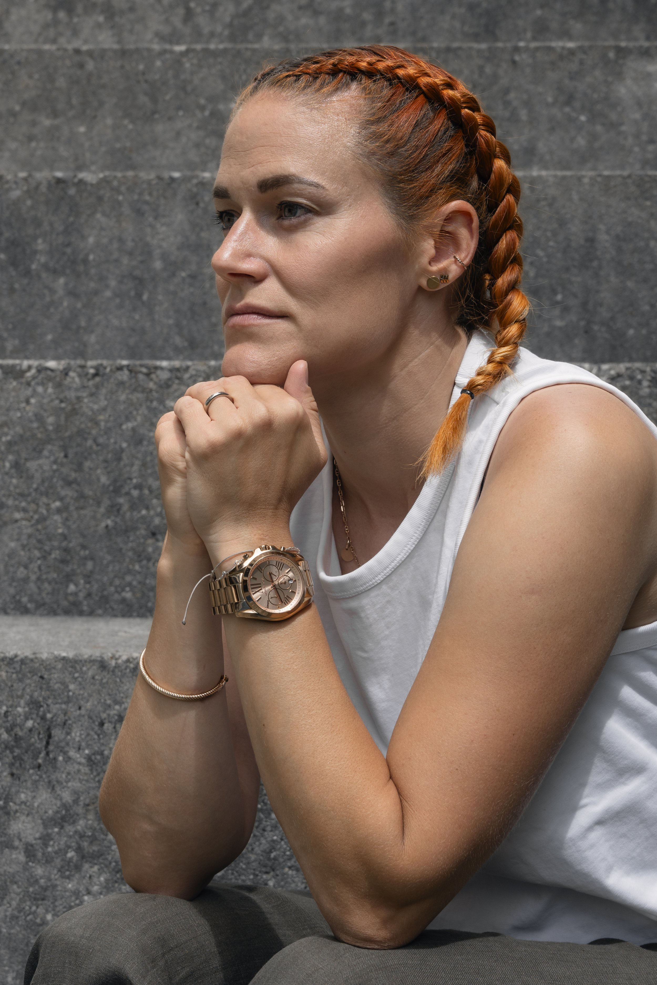 Frau mit roten Zöpfen, Tribals, Sonnenbrille, weißem Tanktop, sitzt auf Stufen, blickt nach rechts, zeigt Handgelenk mit goldener Uhr, trägt Goldschmuck.