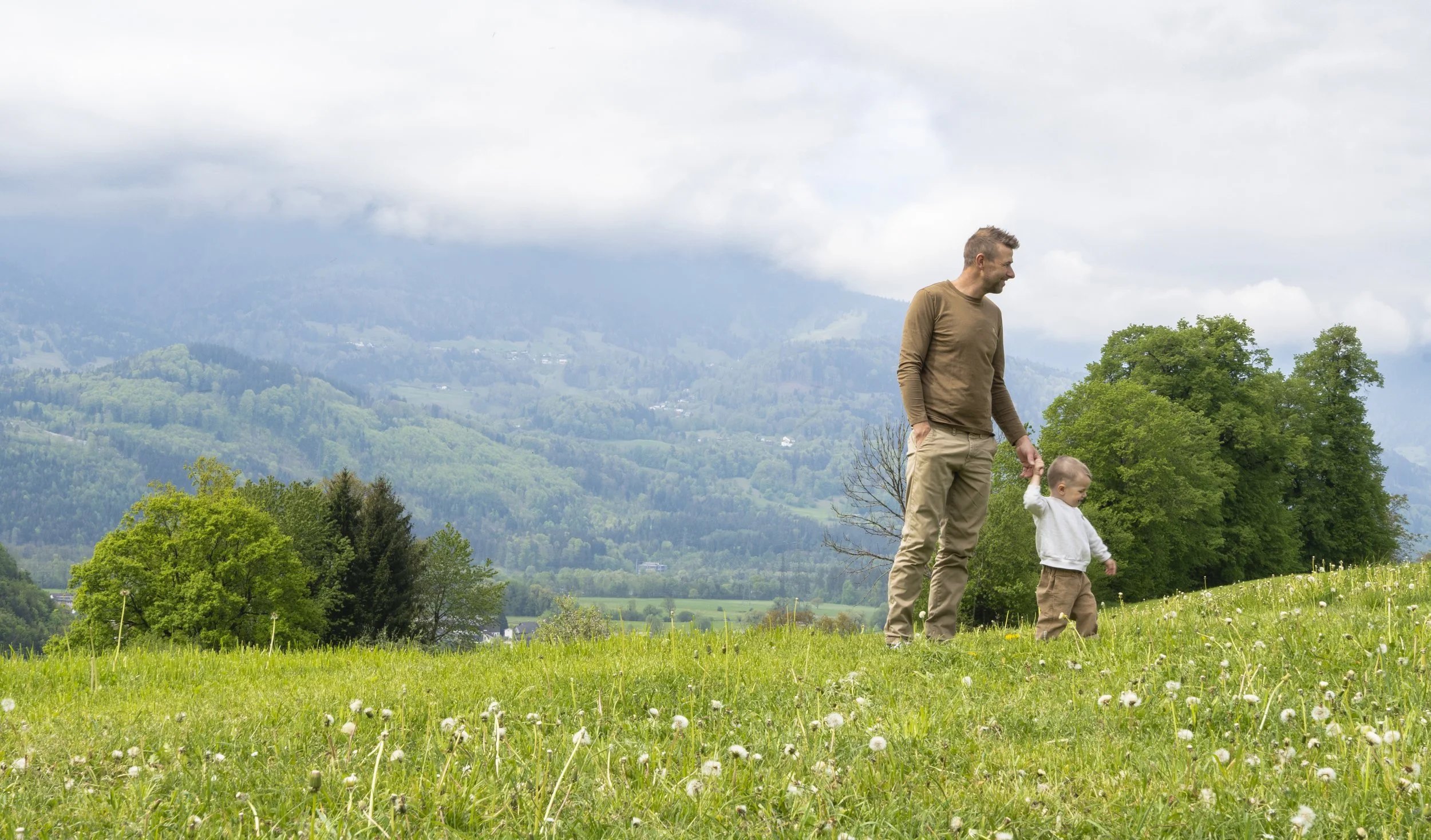 Vater und Kind beim Spaziergang auf einer grünen Wiese mit Bergpanorama im Hintergrund.