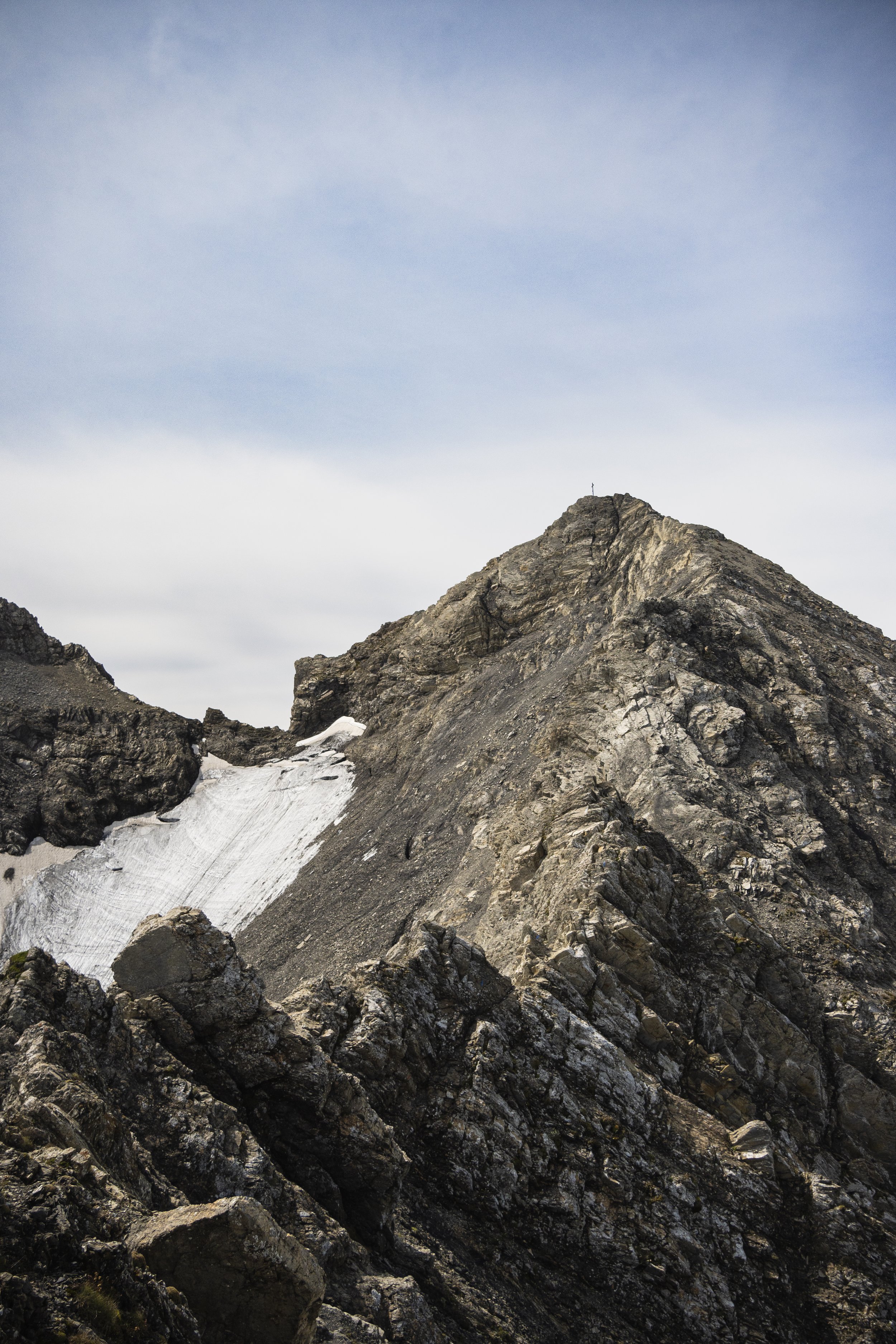 Berggipfel mit Felsen und Schneeresten unter einem bewölkten Himmel.