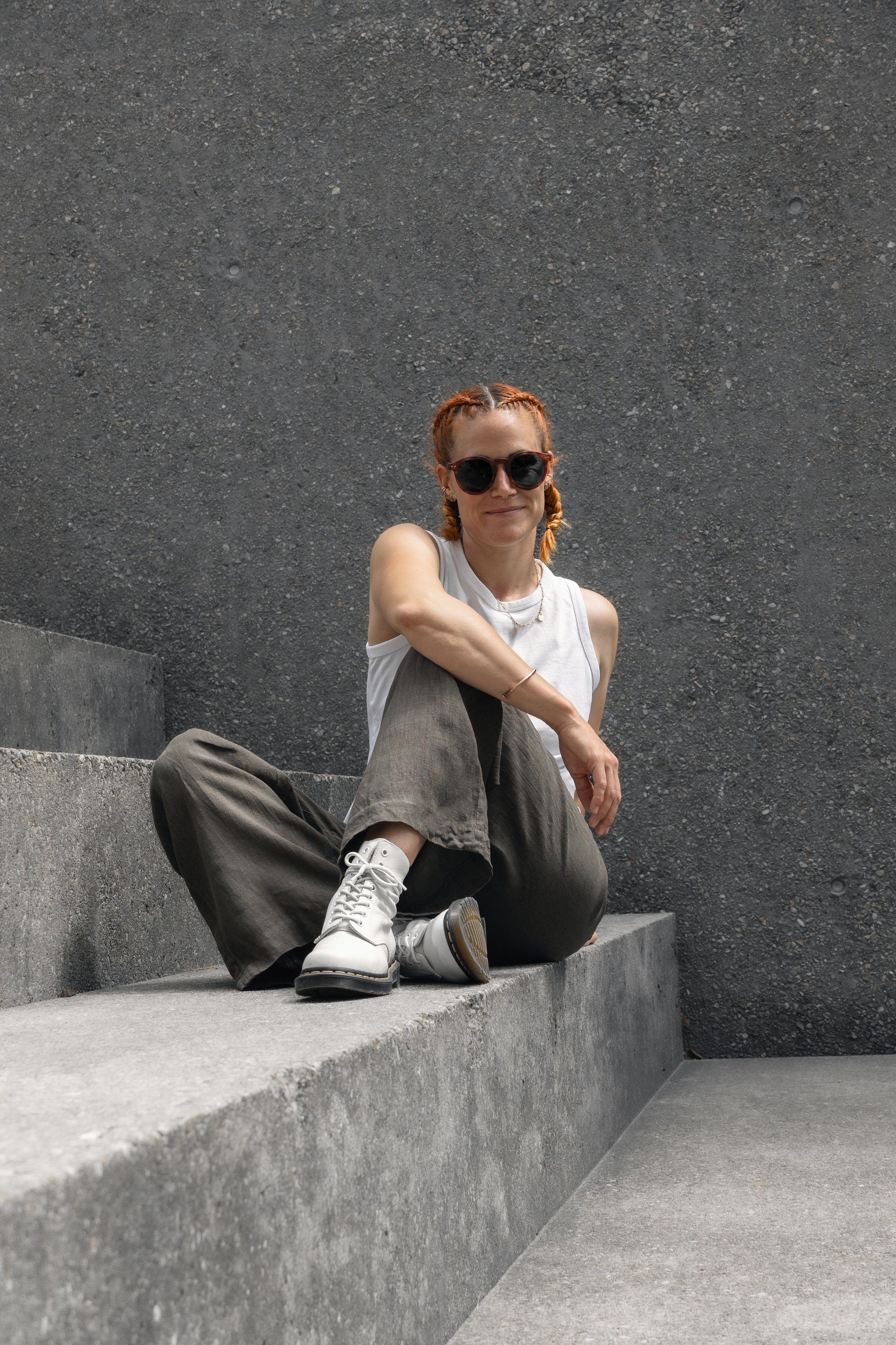 Eine junge Frau mit roten Zöpfen, dunkler Sonnenbrille und weißen Stiefeln sitzt lächelnd auf grauen Betonstufen vor einer dunklen Wand.