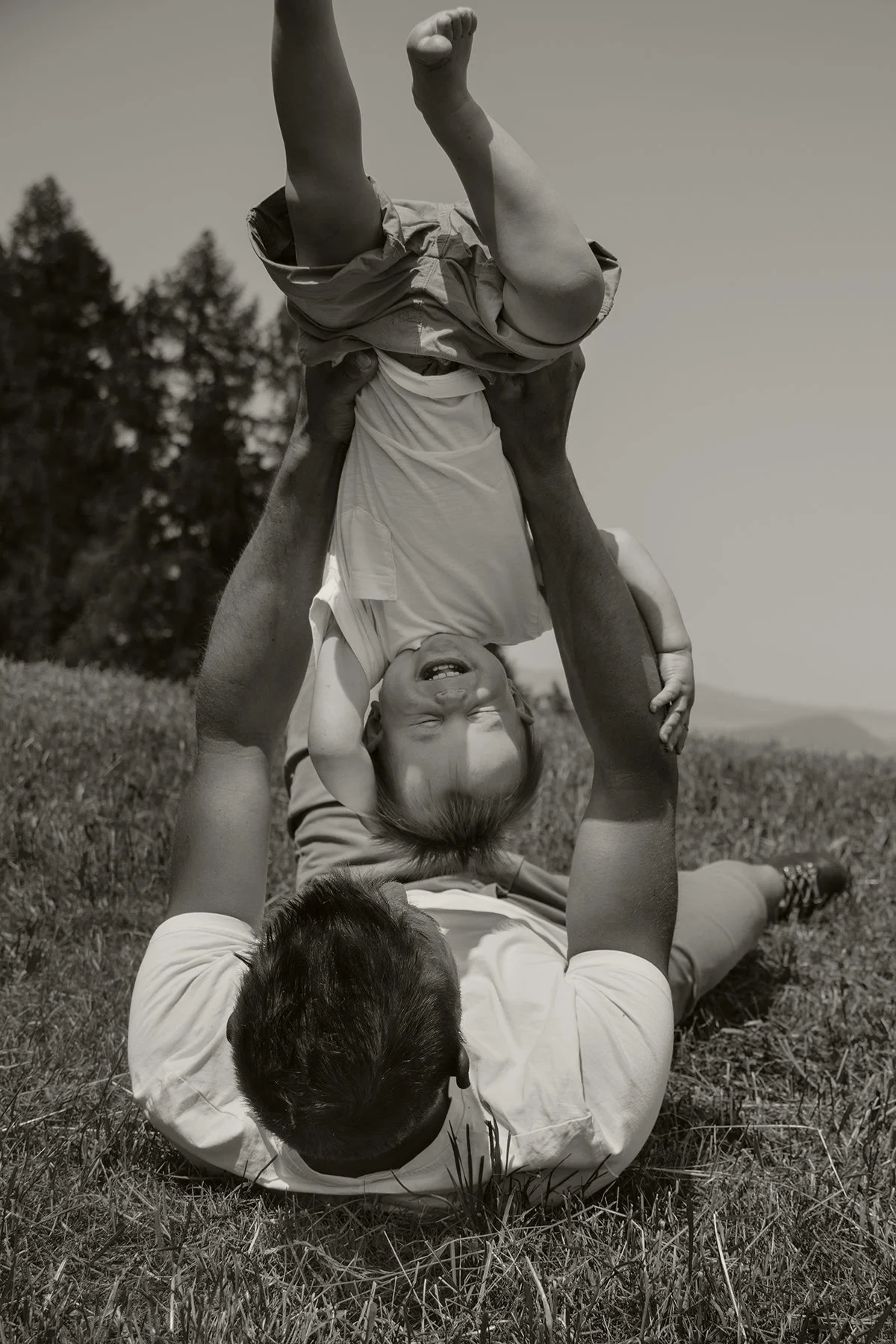 Ein Mann liegt auf dem Boden, hält ein lachendes Kind kopfüber in die Luft, während sie im Freien auf Gras sitzen, mit Bäumen im Hintergrund.