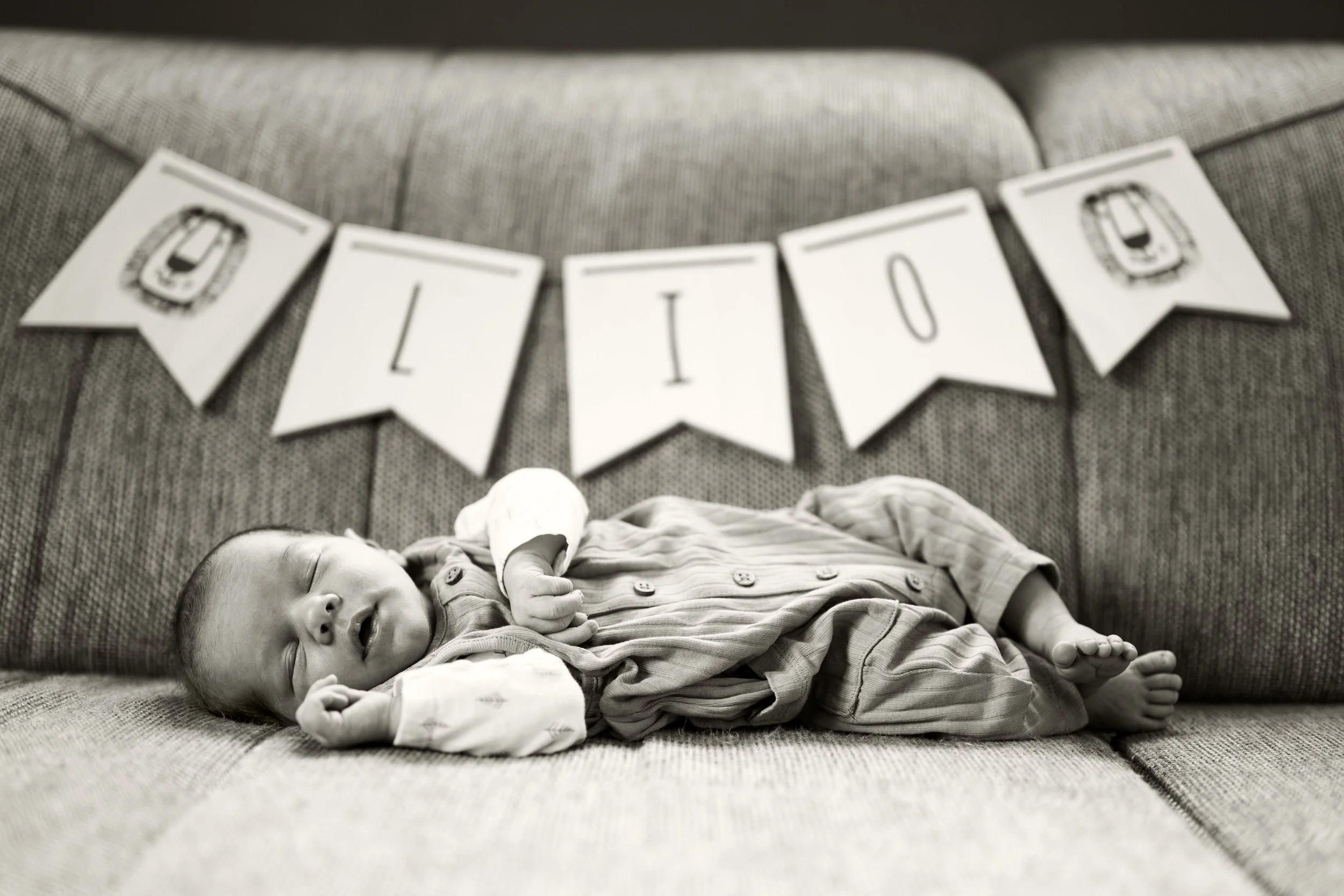Ein Baby liegt gemütlich auf der Couch. Im Hintergrund steht sein Name auf einer Wimpelkette. Lio. Die Aufnahme ist schwarz-weiß.