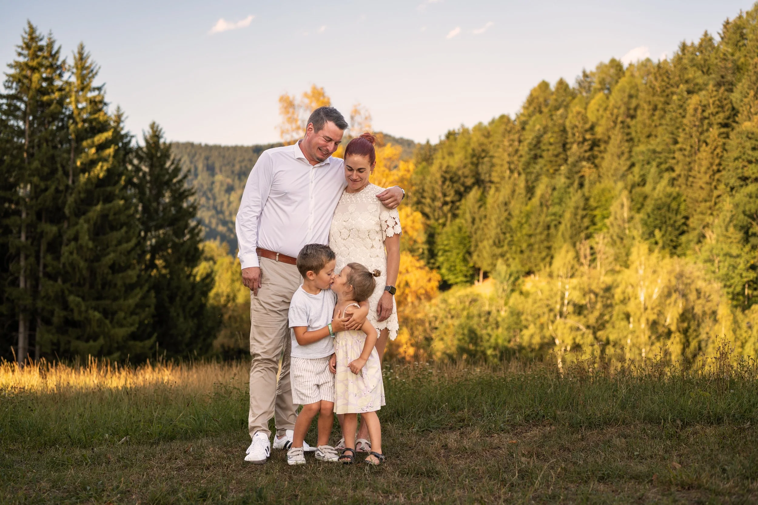 Eine 4-köpfige Familie steht auf einer Wiese, im Hintergrund bunt gefärbte Bäume, das Licht strahlt. Die Eltern stehen hinter ihren Kindern und blicken Lächelnd auf sie. Sie schließen sich in den Arm und Küssen sich.