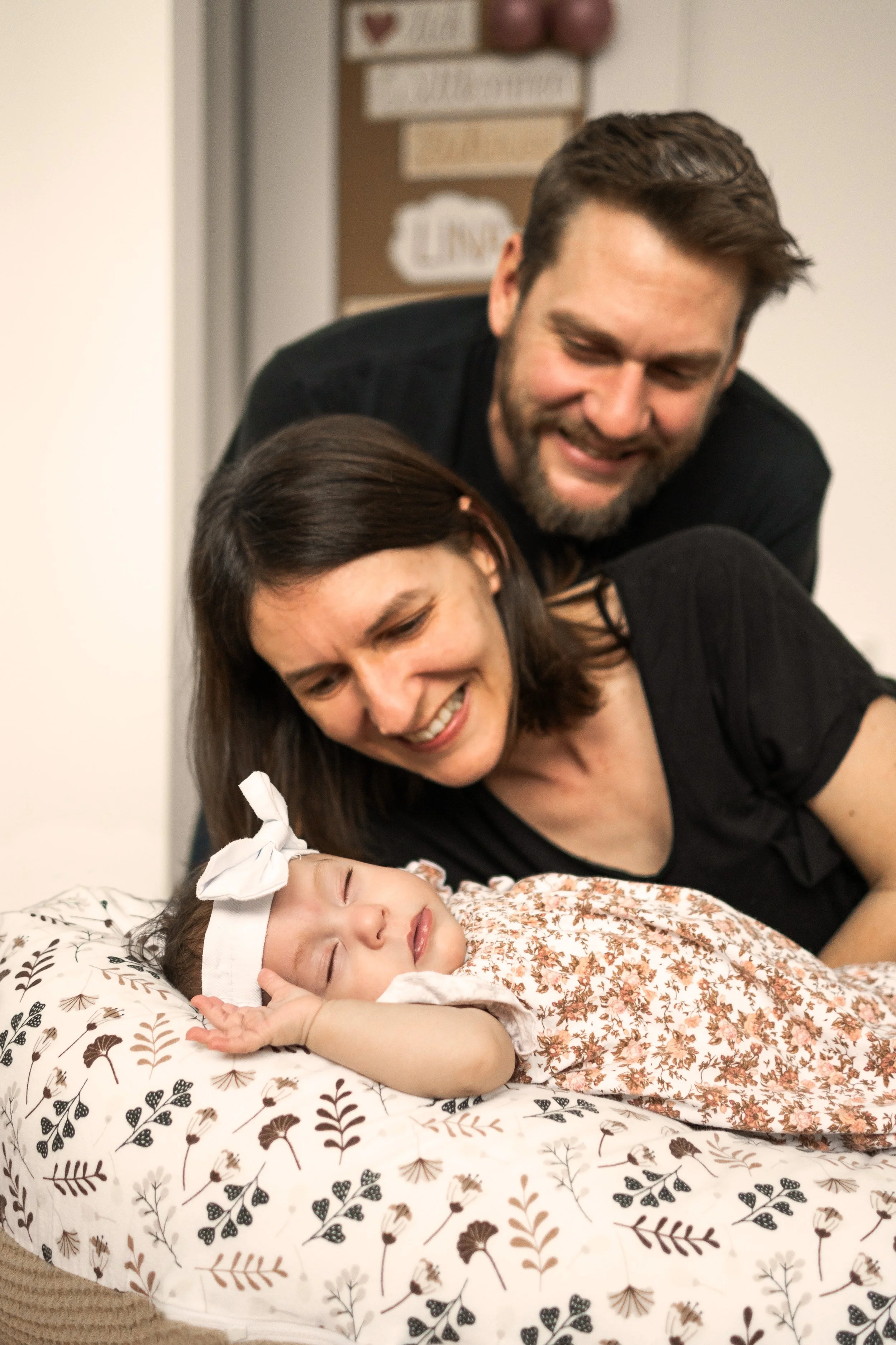 Ein Baby liegt entspannt auf einem Kissen und schläft. Im Hintergrund die glücklichen Eltern.